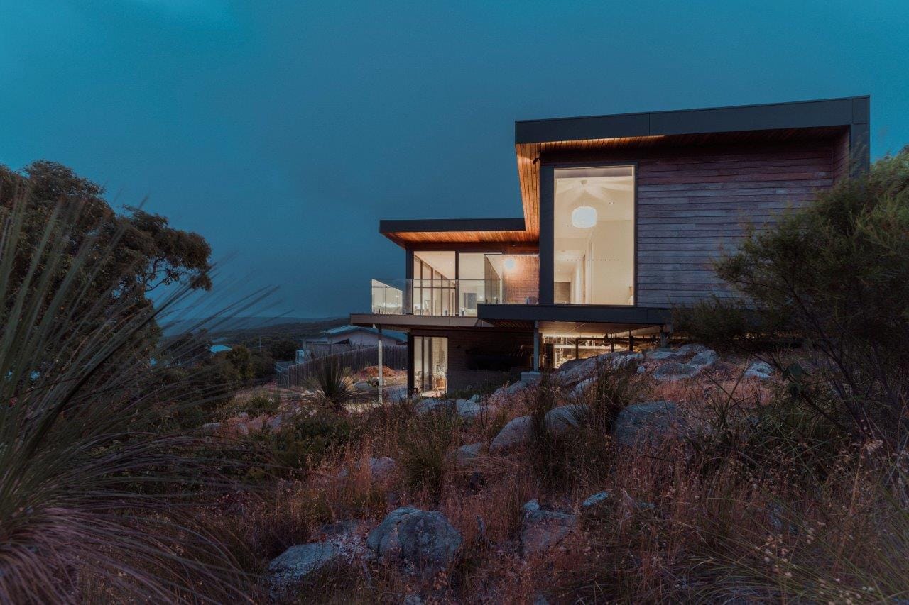 Contemporary timber-clad house illuminated at dusk, set among rocky coastal bushland.