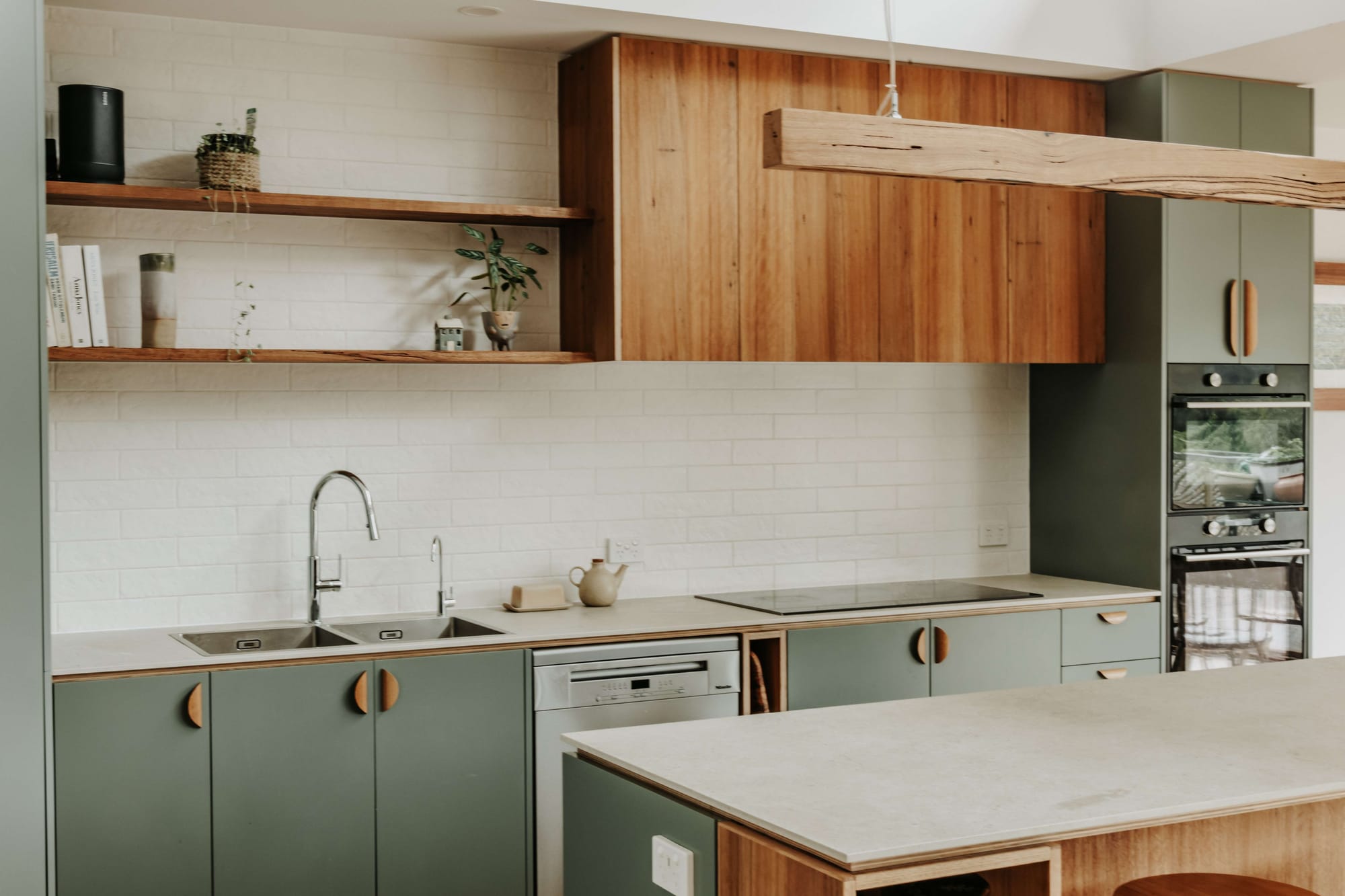 Kitchen with sage-green cabinetry, rounded timber handles, timber overhead cupboards and a suspended timber light above the island.