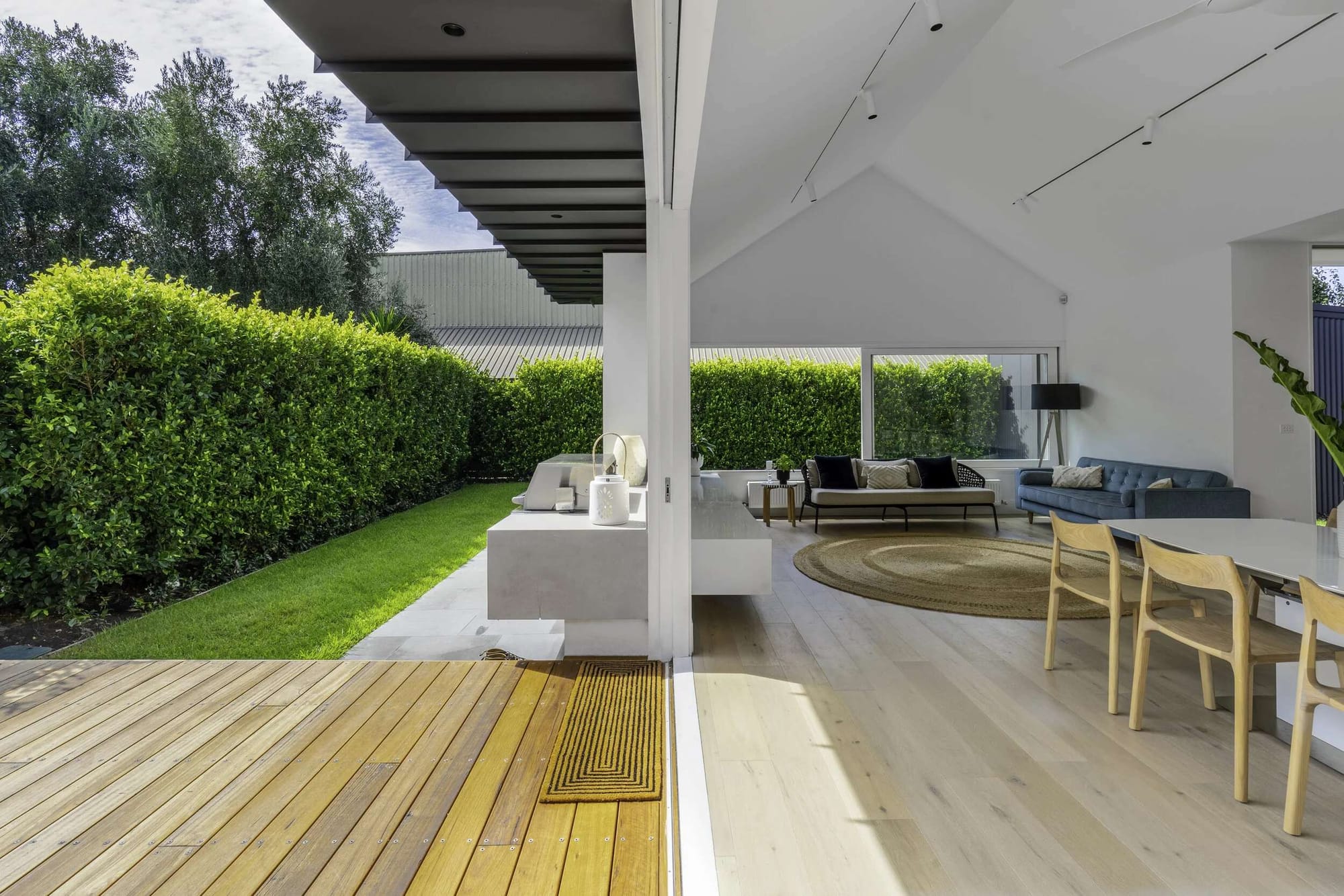 Open living and dining space with a high pitched ceiling and pale timber flooring extending towards a deck and garden at Highett Residential, with interiors by Mikho Interior.