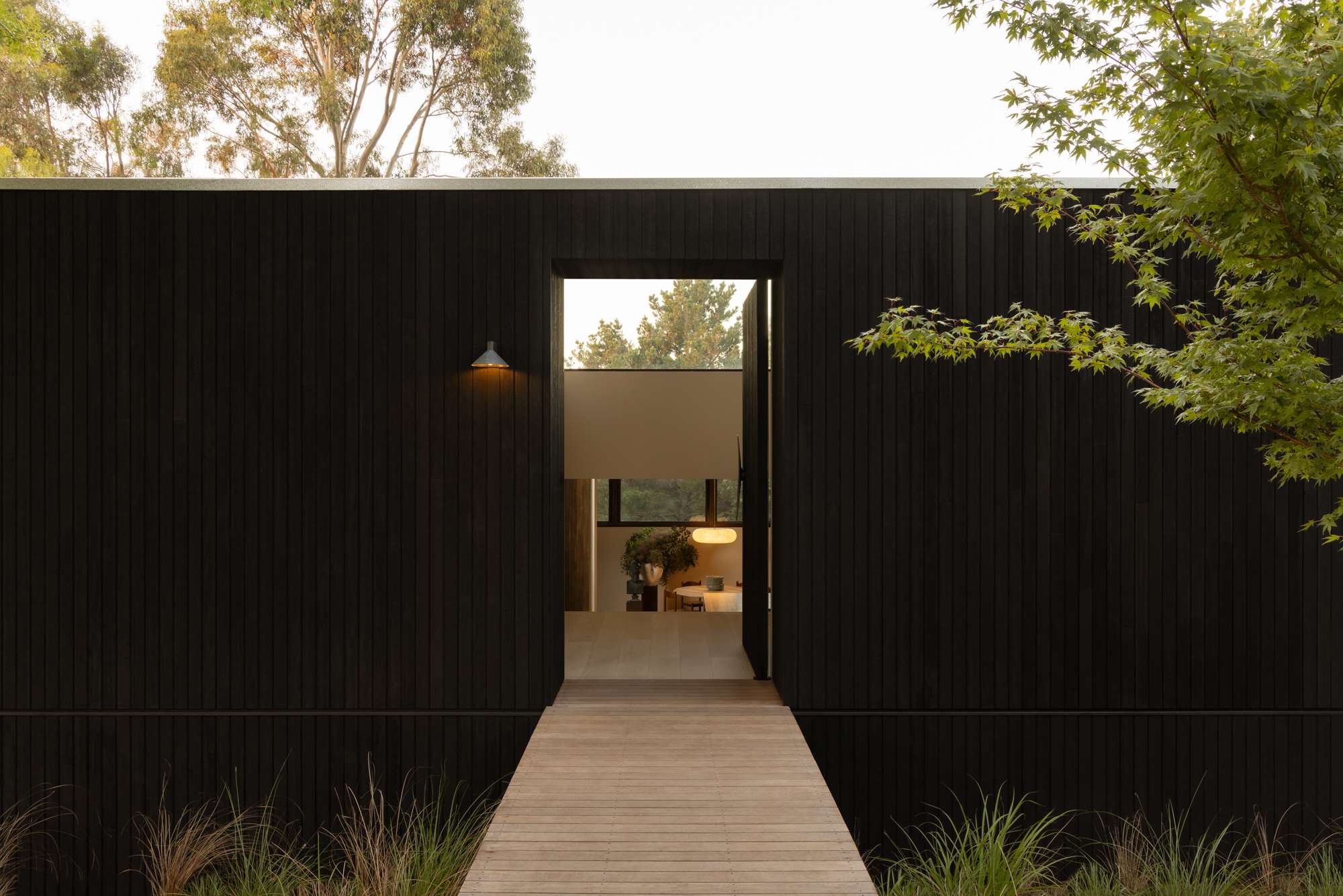 Black timber-clad pavilion with a single doorway opening onto a timber boardwalk through native grasses, lit from within at dusk, by Telha Clarke.
