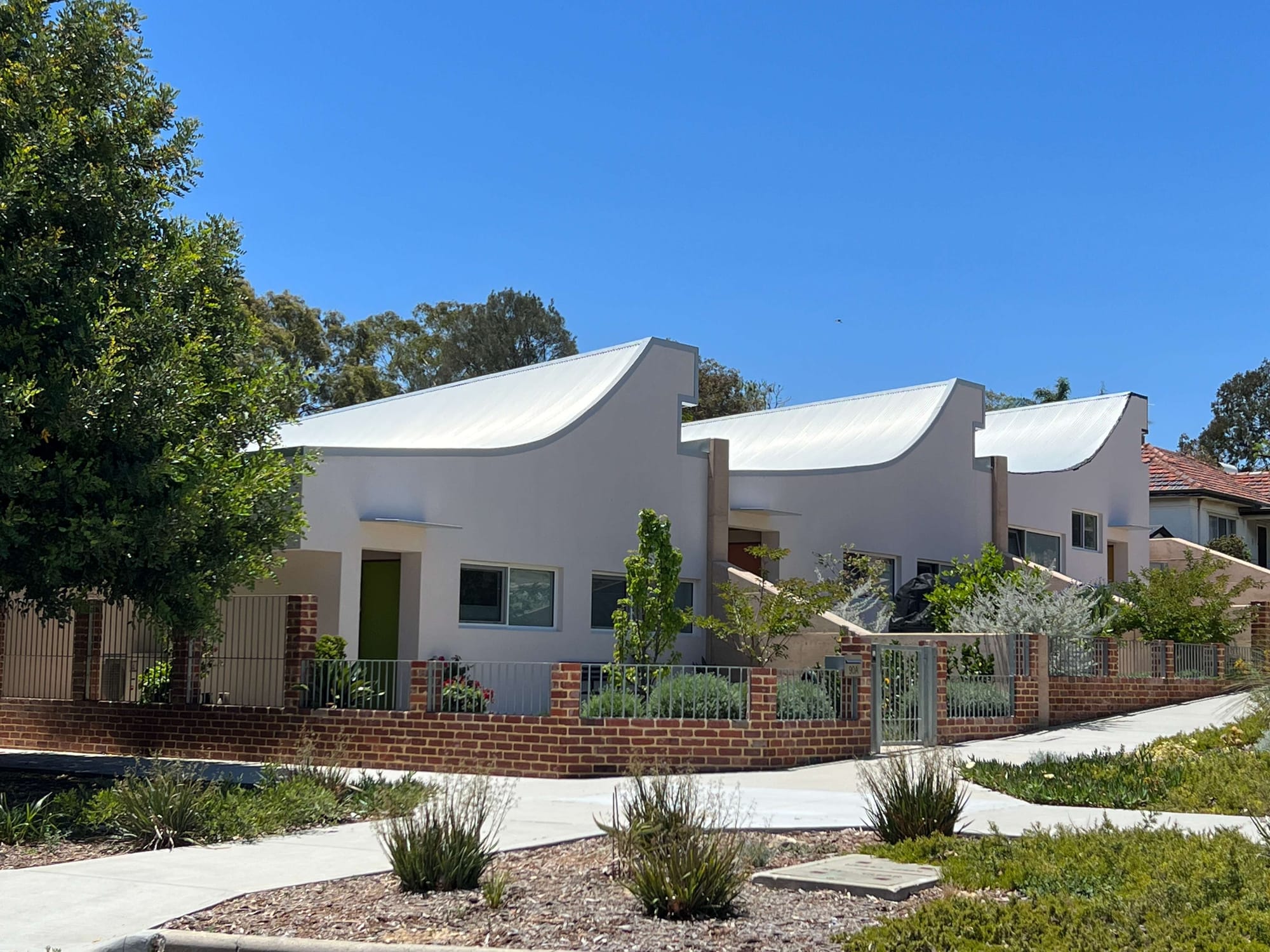 Row of white homes with sweeping curved metal roofs, landscaped front gardens and a low brick-and-metal fence.