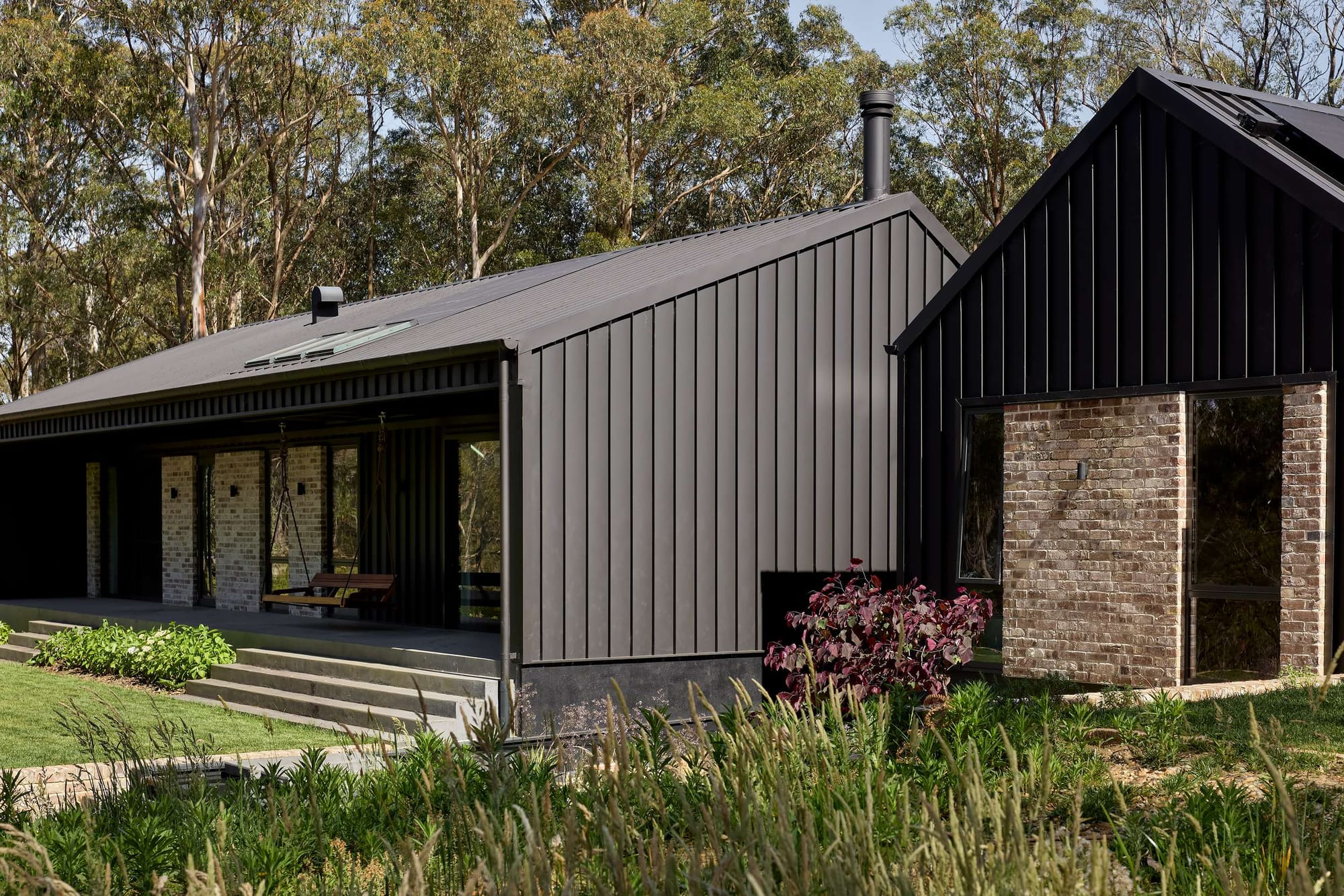 Compact rural cabin with a steep metal roof and large front window, surrounded by grassland, rocks and mature trees. Photography by Claudia Farrar.