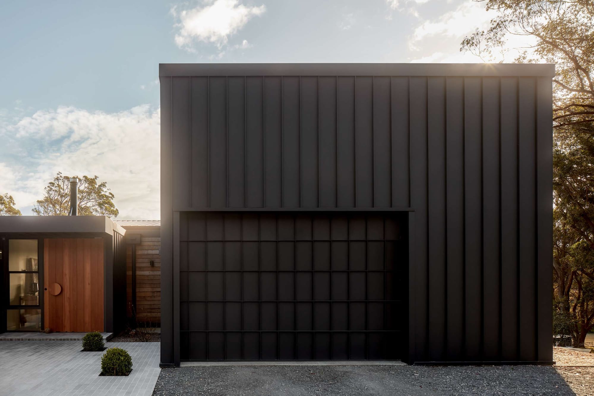 Dark metal-clad garage with a matching panelled door beside a timber entrance and paved pathway.