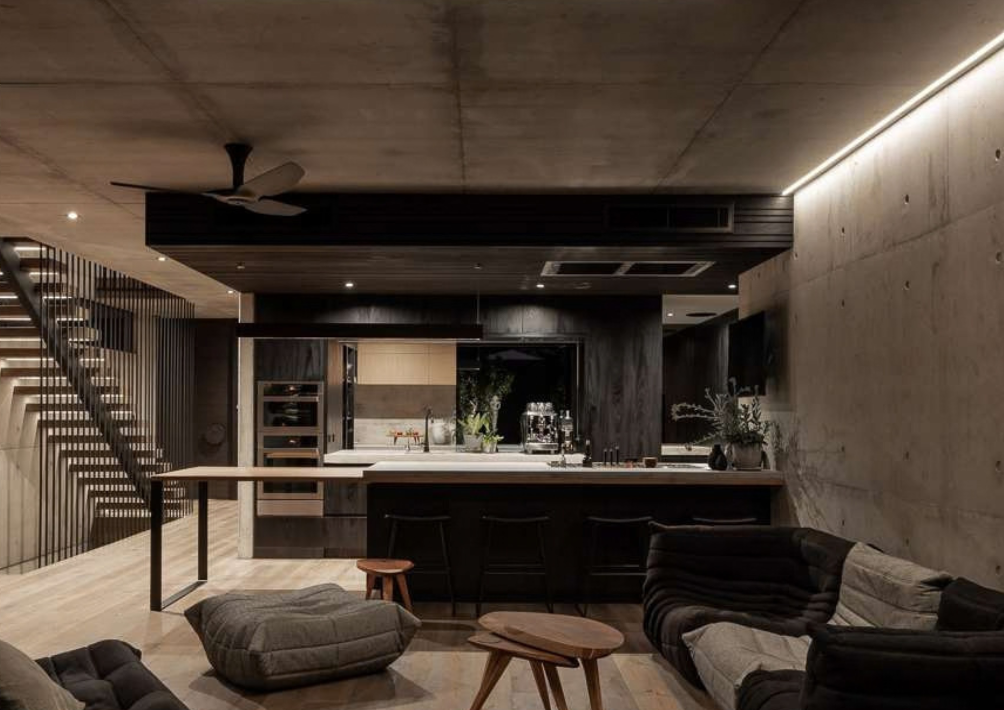 Open kitchen and living space by Marcus Browne Architect. with dark timber joinery, concrete walls, island seating and a staircase screened by black battens.