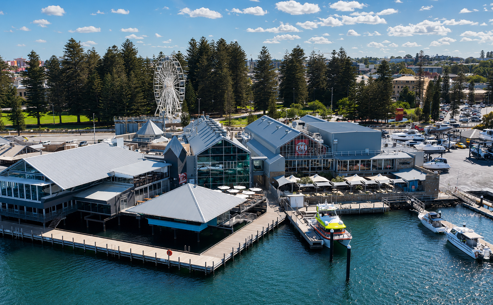 Aerial view of a waterfront entertainment precinct and marina, with a Ferris wheel and tall pine trees beyond.