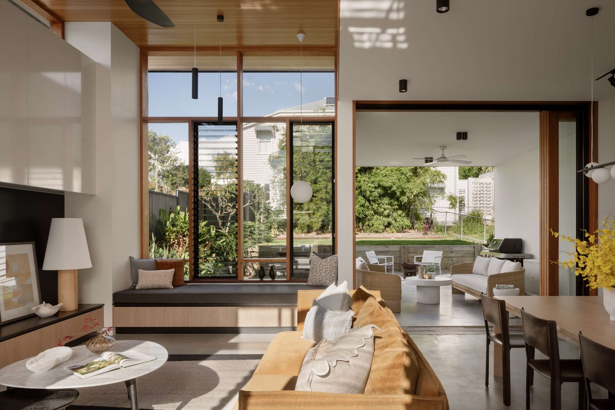 Sunlit living room with a built in window seat, tall louvre windows and a timber ceiling opening to a covered terrace at Light Pavilion House by FOLAR Architecture & Interiors.