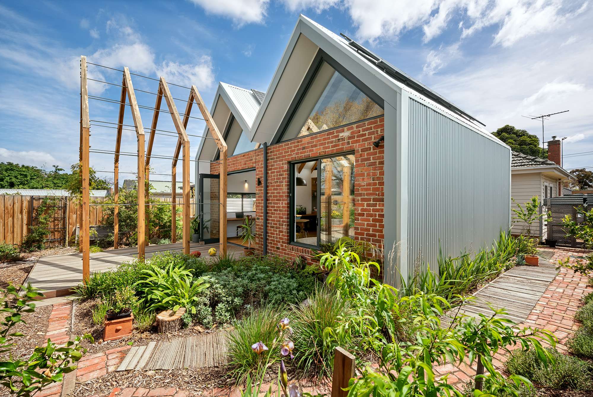 Garden view of Maaemo by Green Sheep Collective, showing a brick and corrugated metal extension with pitched roofs, large windows and a timber pergola.