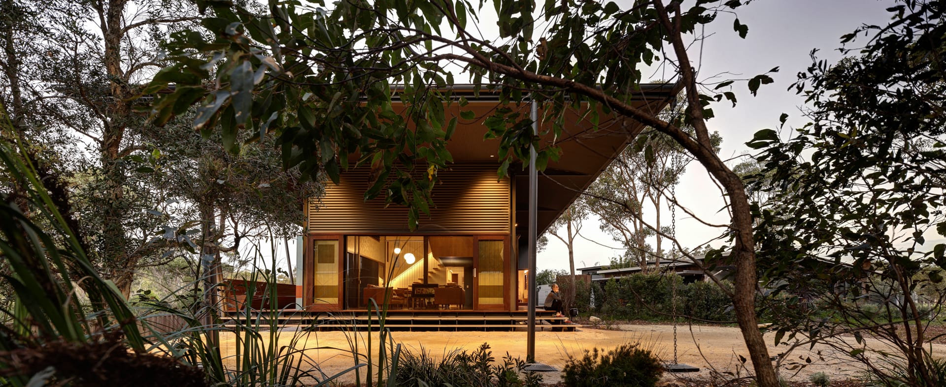 Home by Marra + Yeh with a broad sloping roof, an open timber deck and a warmly lit interior framed by garden trees at dusk.