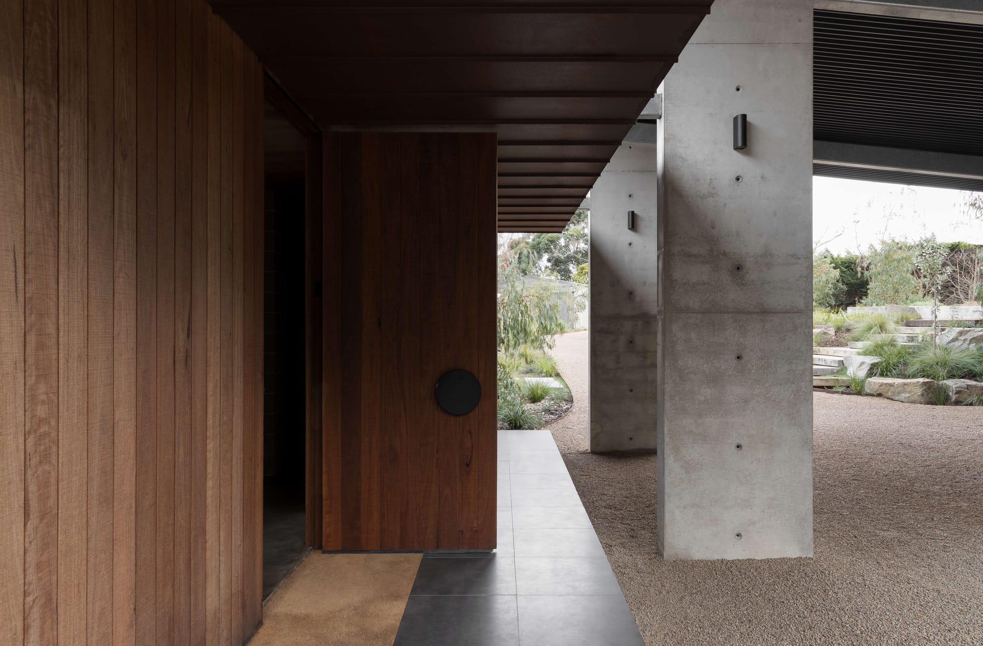 Timber entrance door with a round black handle beside exposed concrete columns and a sheltered garden walkway.