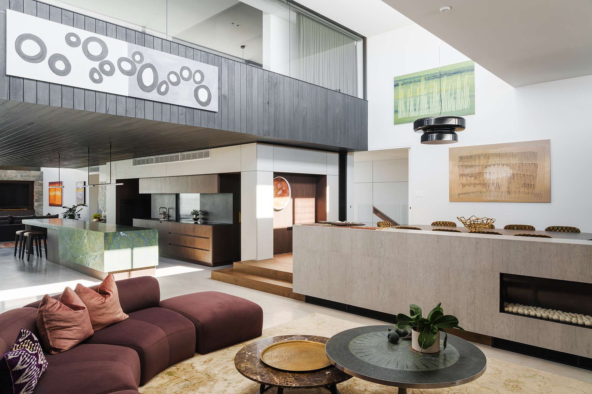 Sunlit living area beneath a glazed mezzanine, with a curved burgundy sofa, stone fireplace divider and green kitchen island at Mezzanine House by CSA Craig Steere Architects.