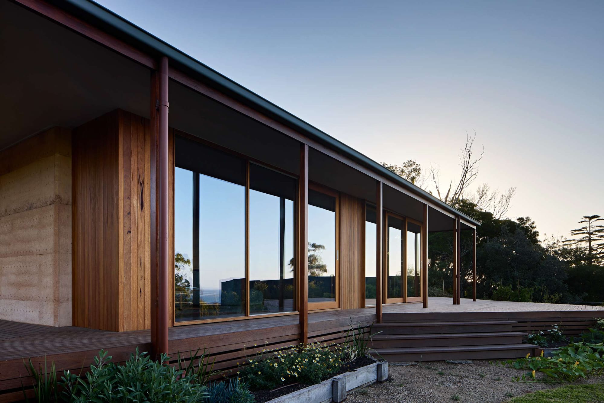 Long rammed-earth and timber house with a deep verandah and floor-to-ceiling glazing along a timber deck, photographed at dusk, by FMD Architects.