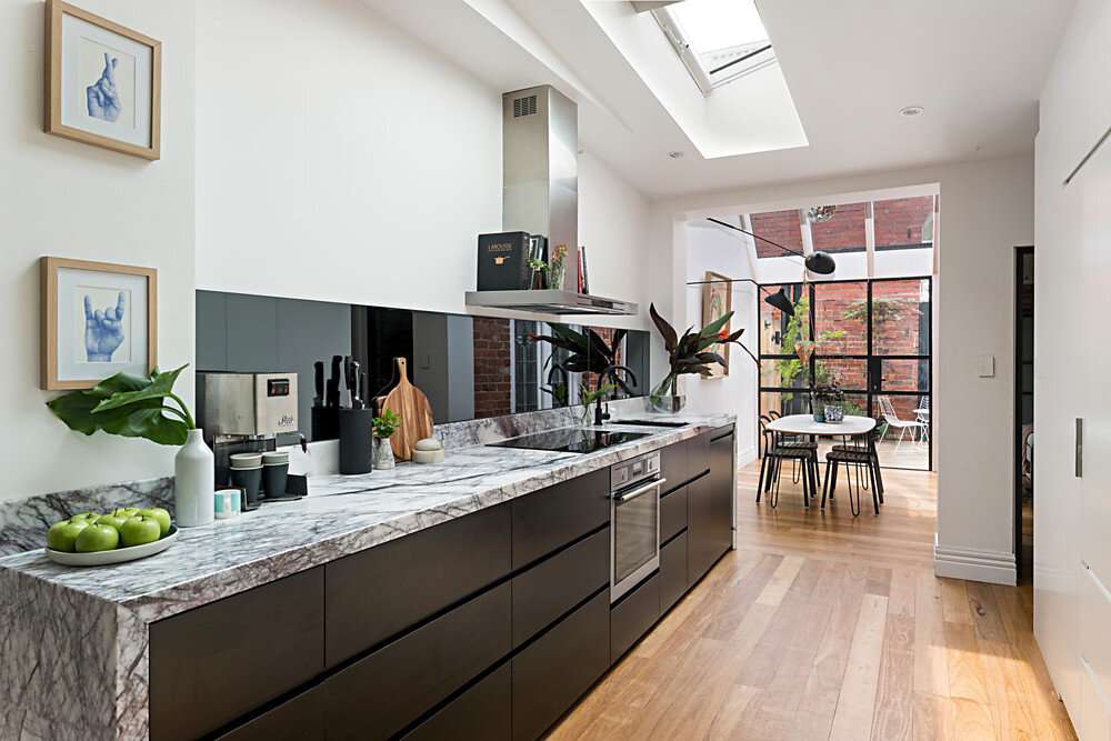Skylit galley kitchen with dark cabinetry, a veined stone benchtop and timber flooring leading to a glazed dining area at Northcote Residence by Colab Architects.