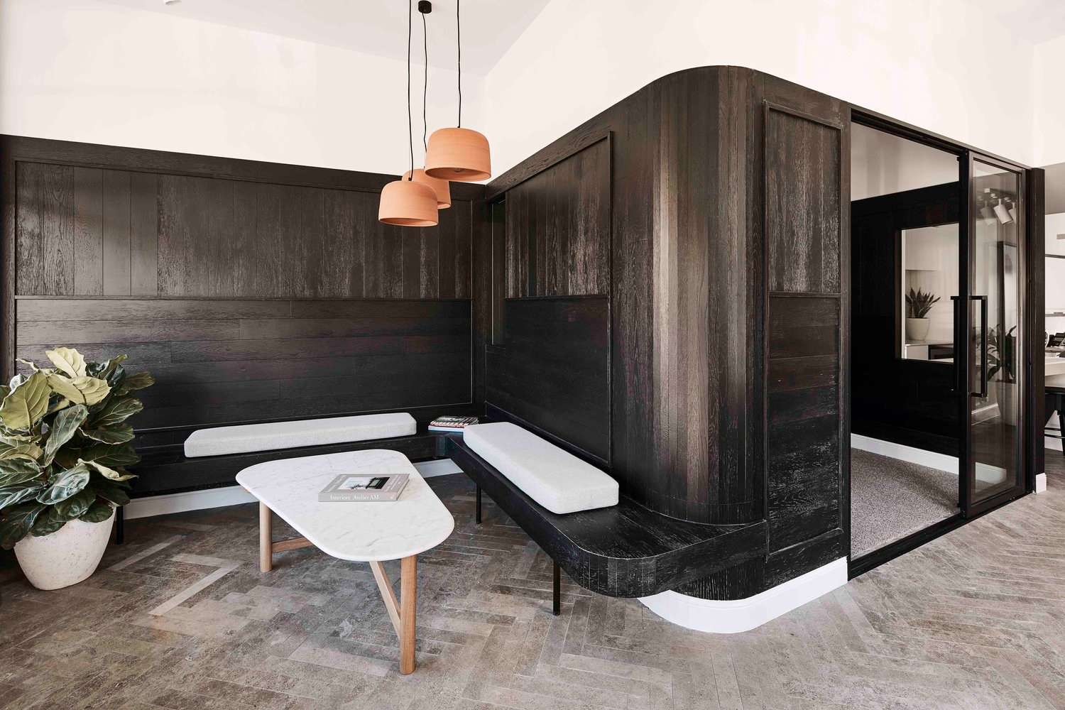 Office meeting nook with curved dark timber joinery, upholstered bench seating, terracotta pendant lights and a glass-walled room.