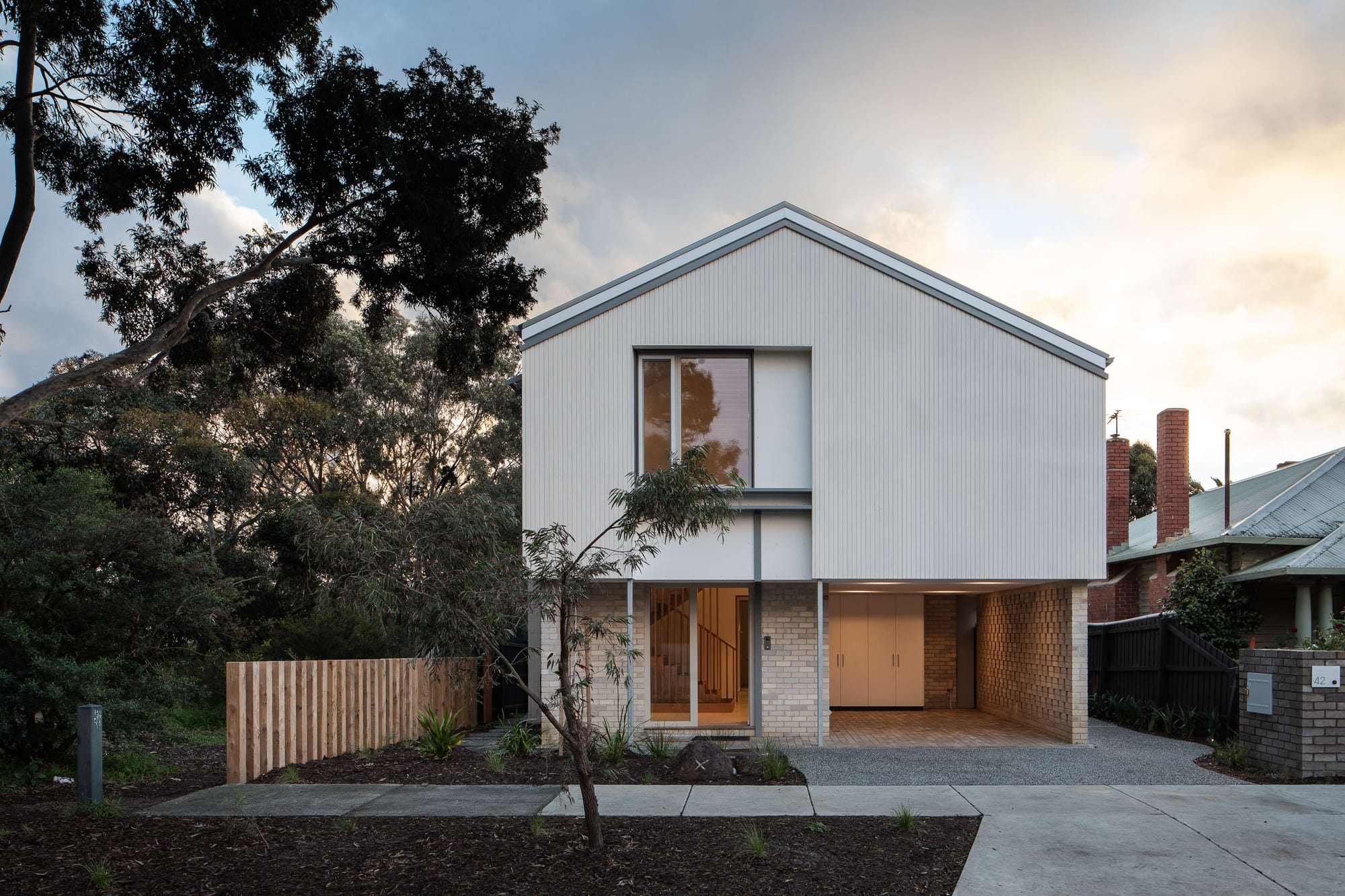 Two-storey home at dusk with a white gabled upper floor, pale brick ground floor and recessed carport framed by trees.