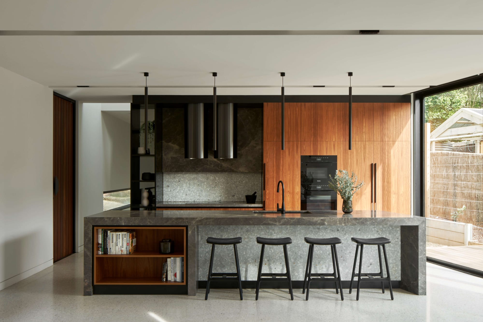 Kitchen with timber cabinetry, a grey stone island, inset bookshelves and four black stools beside large windows at Red Hill Residence by Karavasil Architects.