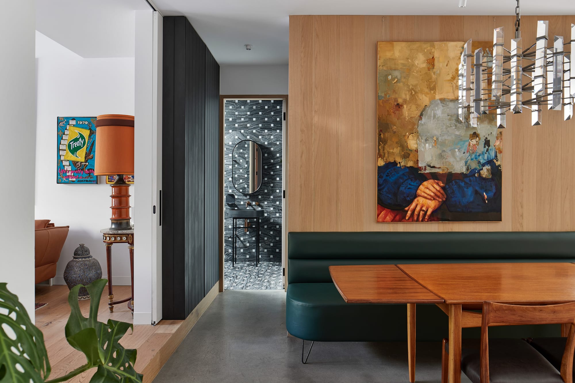 Dining area with a dark green banquette, timber table, colourful artwork and a geometric pendant light, beside a patterned powder room.