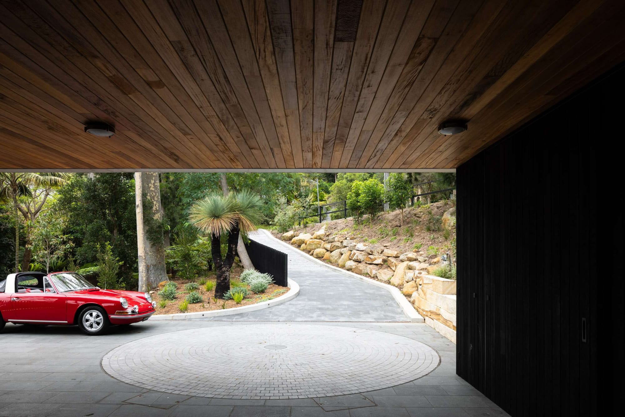 Timber-lined carport with a circular vehicle turntable, red sports car and sloping driveway bordered by stone retaining walls.