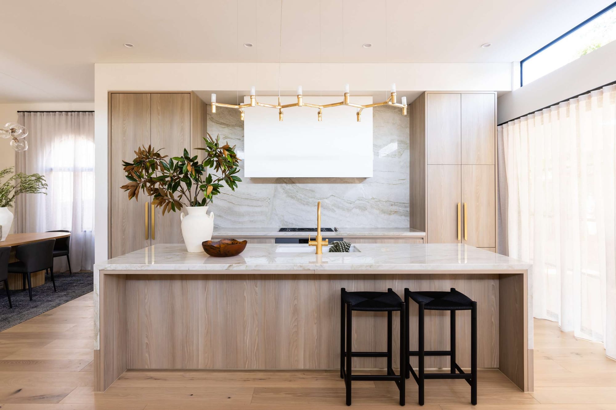 Light filled kitchen by Studio Belle with pale timber cabinetry, a broad stone island, black stools and an adjoining dining space.