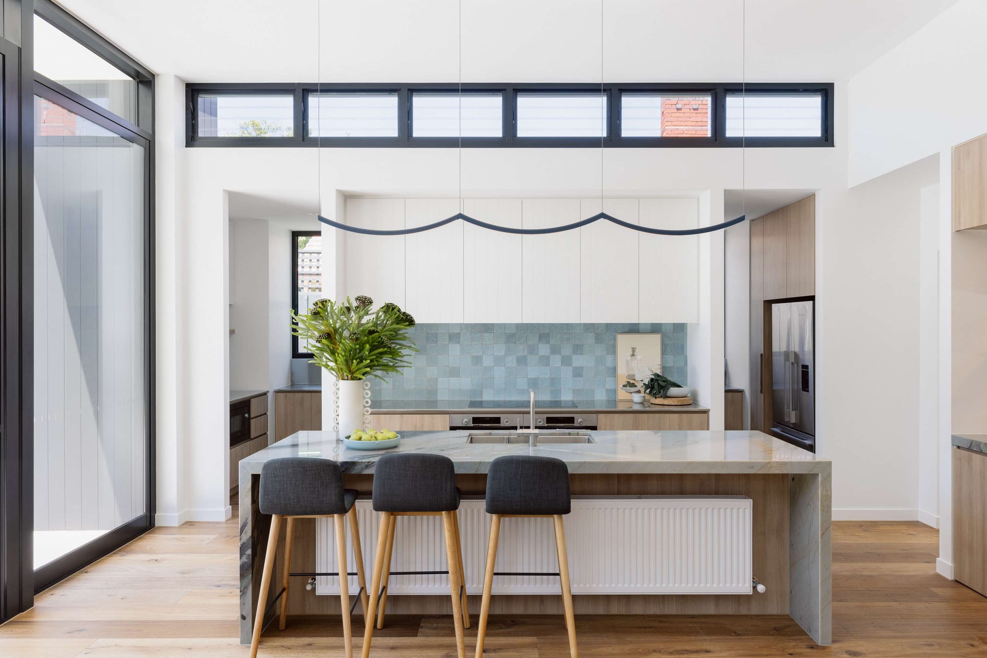 Bright kitchen at Saint George by Flowe Interiors with pale blue tiled splashback, timber cabinetry, stone island and high clerestory windows.
