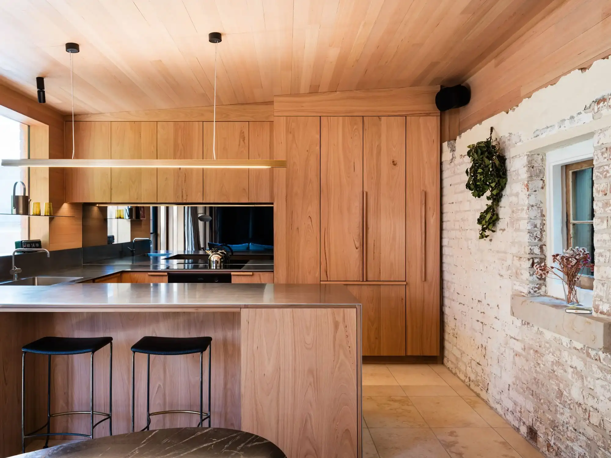 Timber lined kitchen at Sandy Bay Heritage Cottage by Matt Williams Architects, with warm cabinetry, a metal benchtop and the original painted brick wall exposed.
