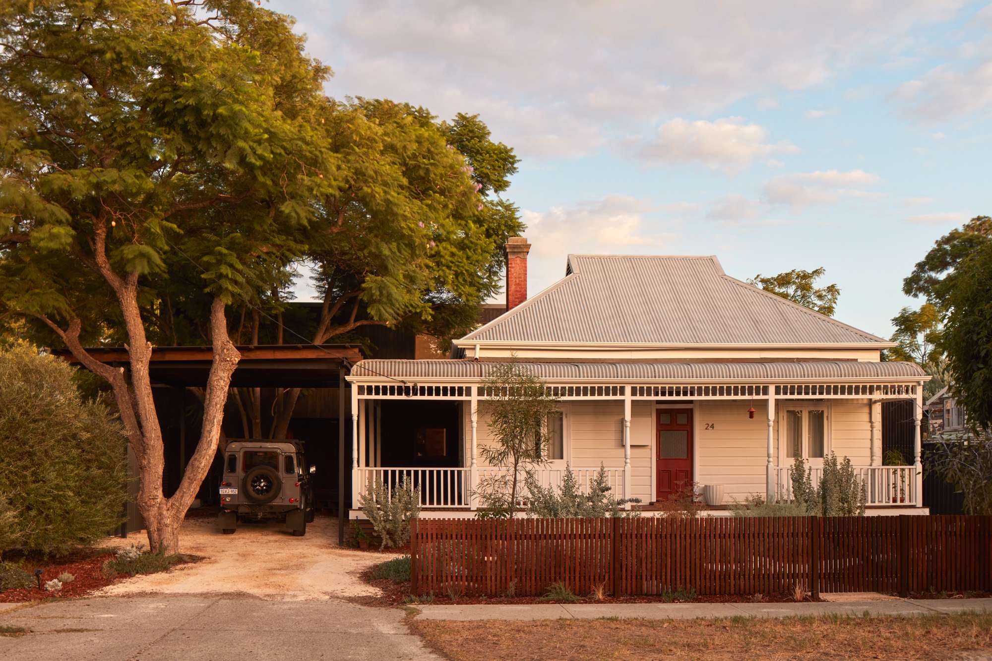 White weatherboard home with a red front door, covered verandah and adjoining carport beneath mature trees.