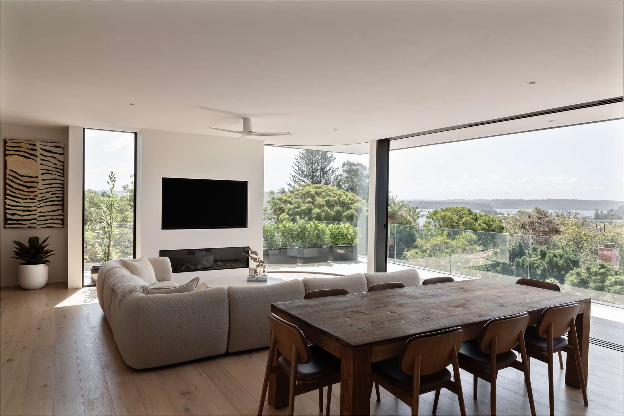 Open living and dining area with timber flooring, a curved cream sofa and expansive glazing overlooking trees and the bay at Stonemason’s House by Spicer Architecture.