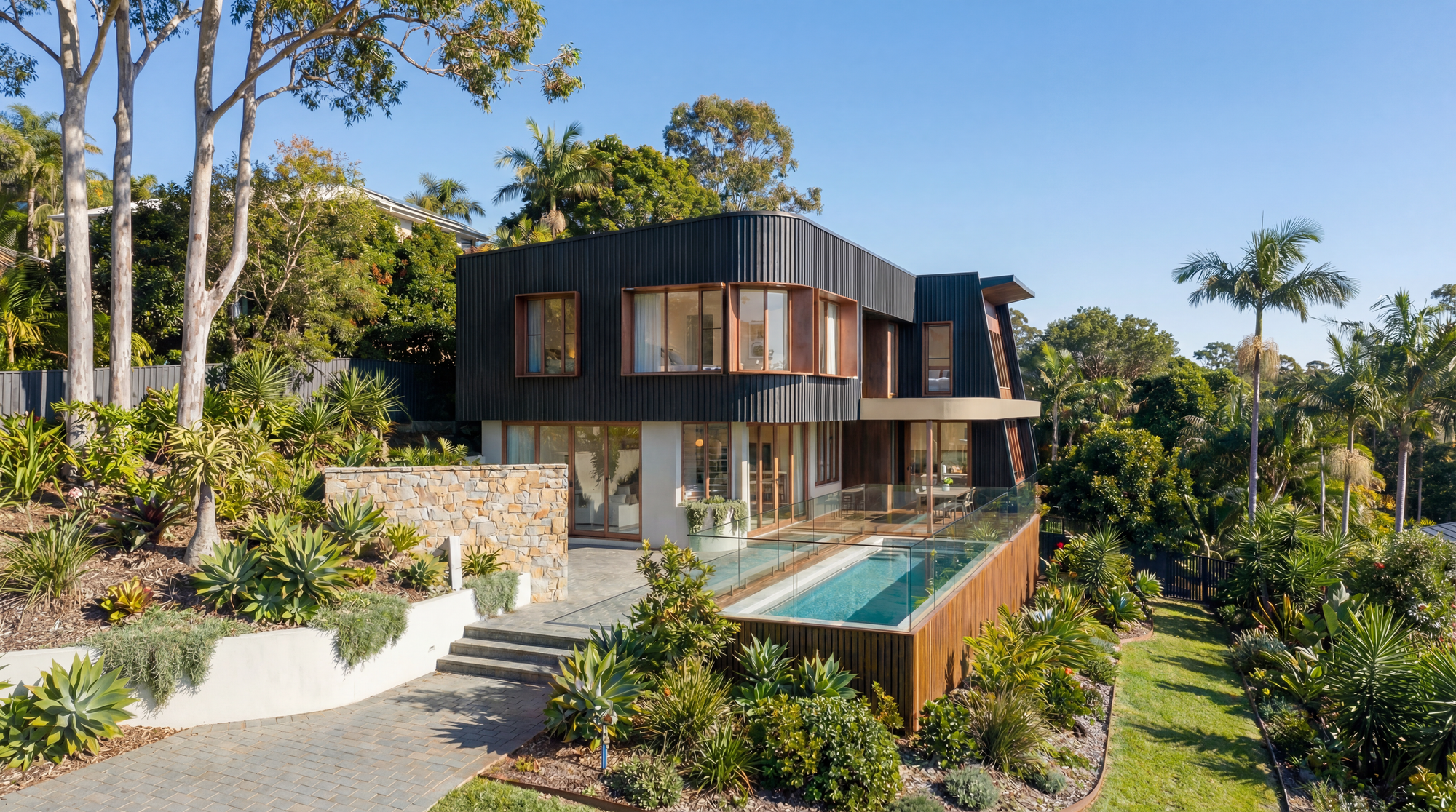 Two-storey home with dark vertical cladding, timber-framed corner windows and a narrow swimming pool set within lush hillside gardens.