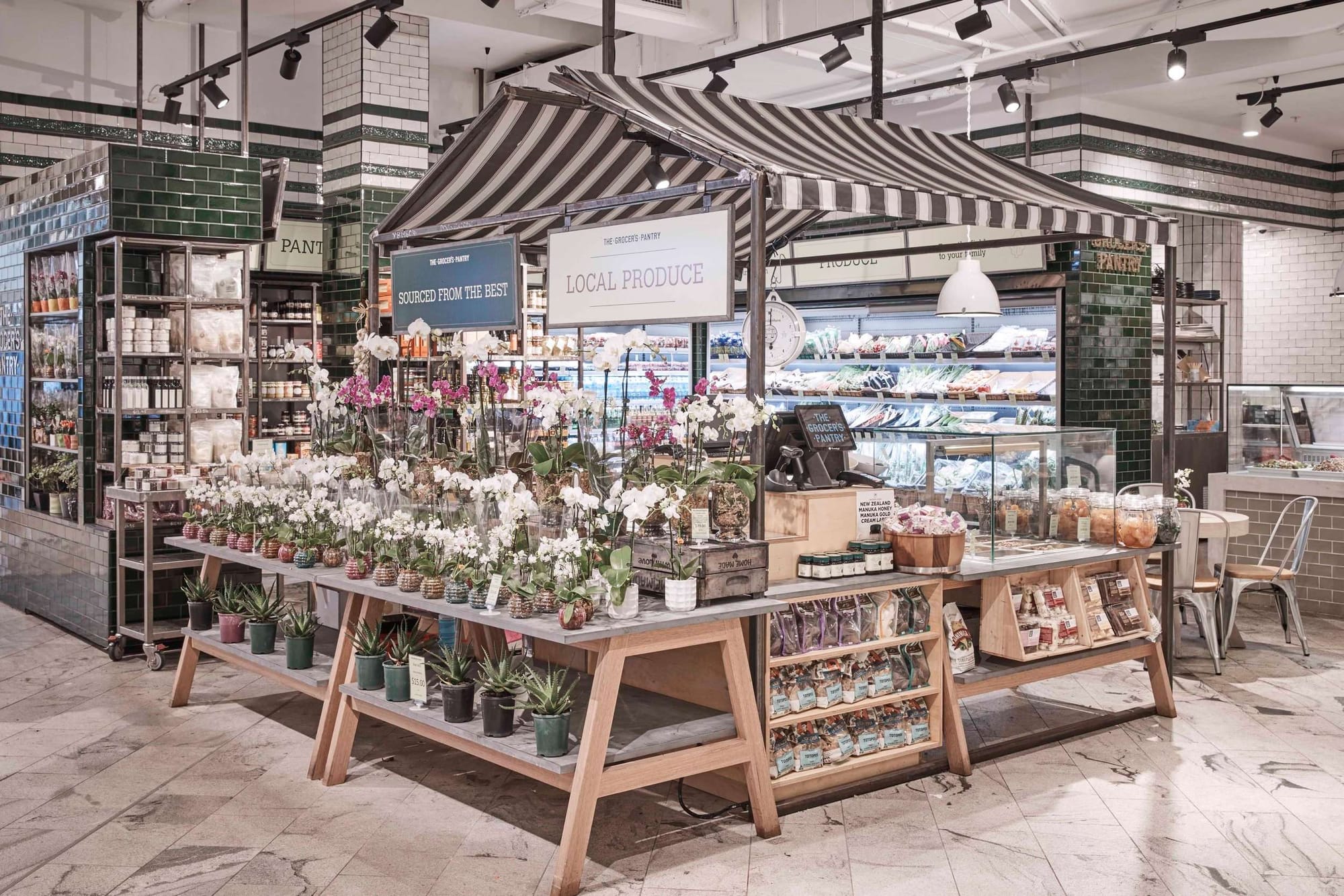 Grocer’s Pantry interior with green and white tiled walls, timber displays, a striped canopy, fresh produce and potted orchids.