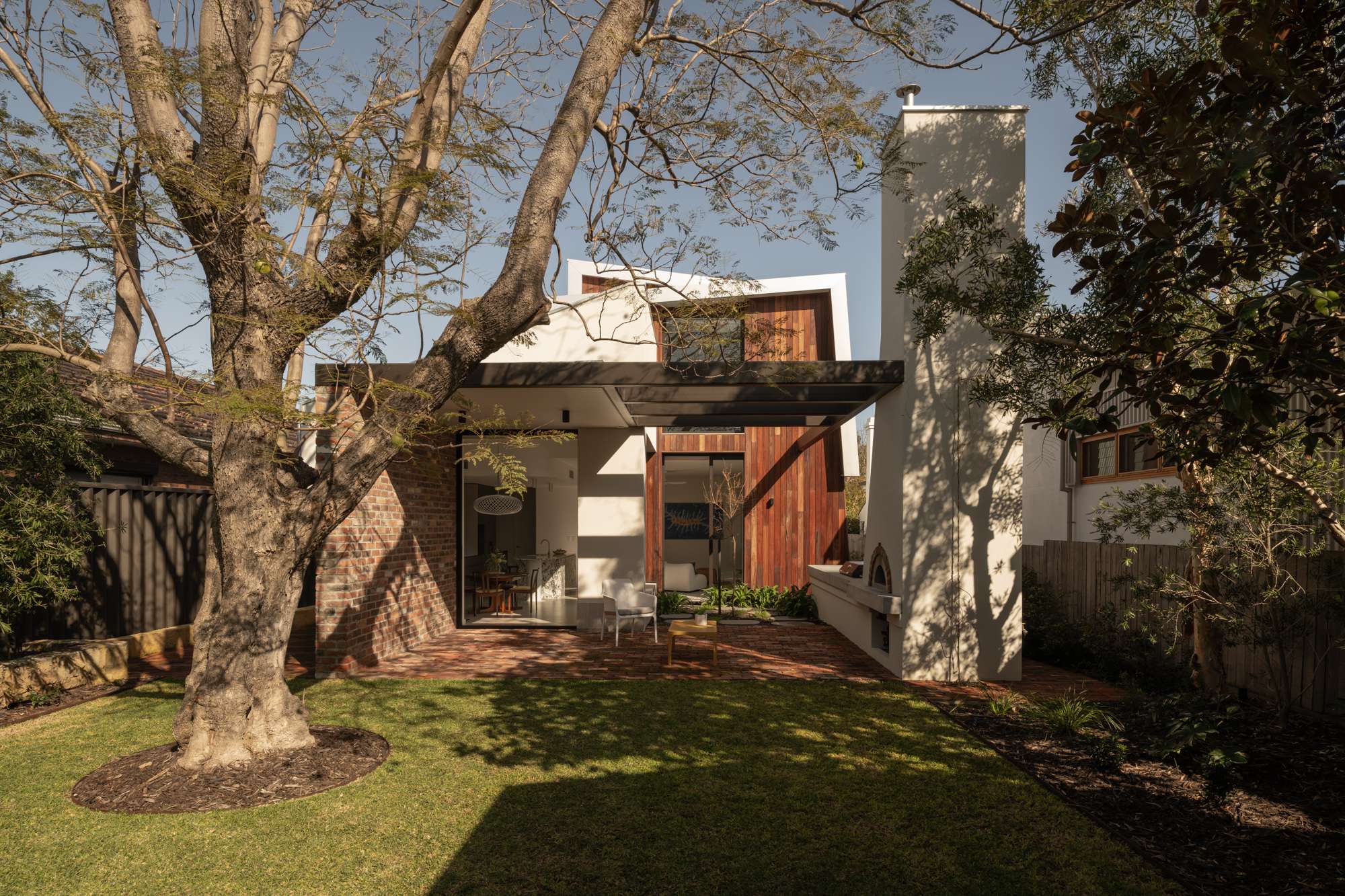 Rear extension to a brick heritage home, with a rendered contemporary addition and chimney form behind a mature tree in the garden, by &Dalecki.