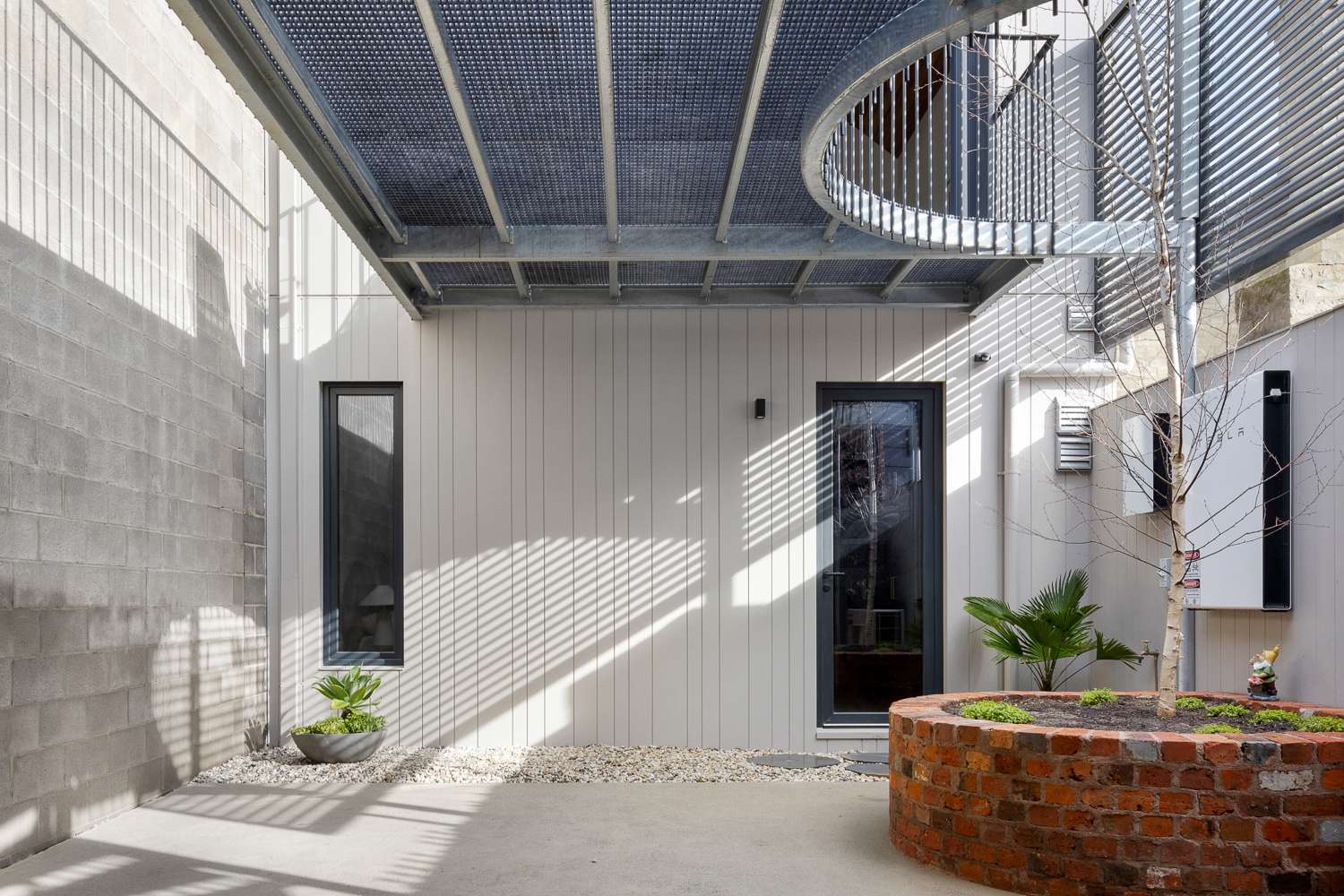 Sunlit courtyard with narrow windows, a circular brick planter and an overhead steel walkway with an opening for a tree.