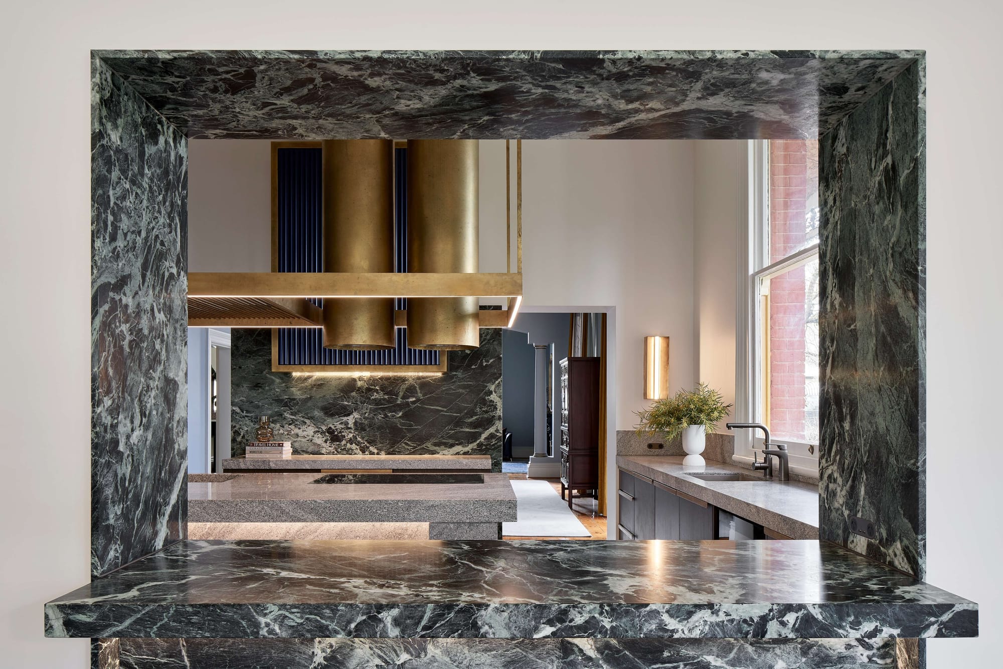 Kitchen by Atelier Bond viewed through a dramatic dark stone opening, with matching stone surfaces, brass rangehoods and daylight from a side window.