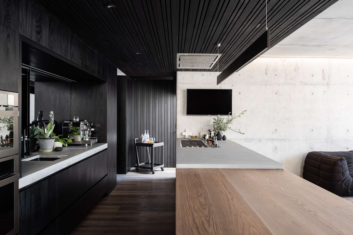 Dark timber kitchen by Marcus Browne Architect. with a concrete island, timber dining surface and exposed concrete wall connecting to the living area.