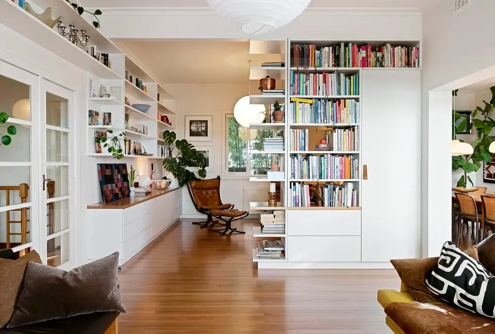 Light-filled living and reading area with white built-in bookshelves, timber flooring, indoor plants and a brown leather lounge chair.