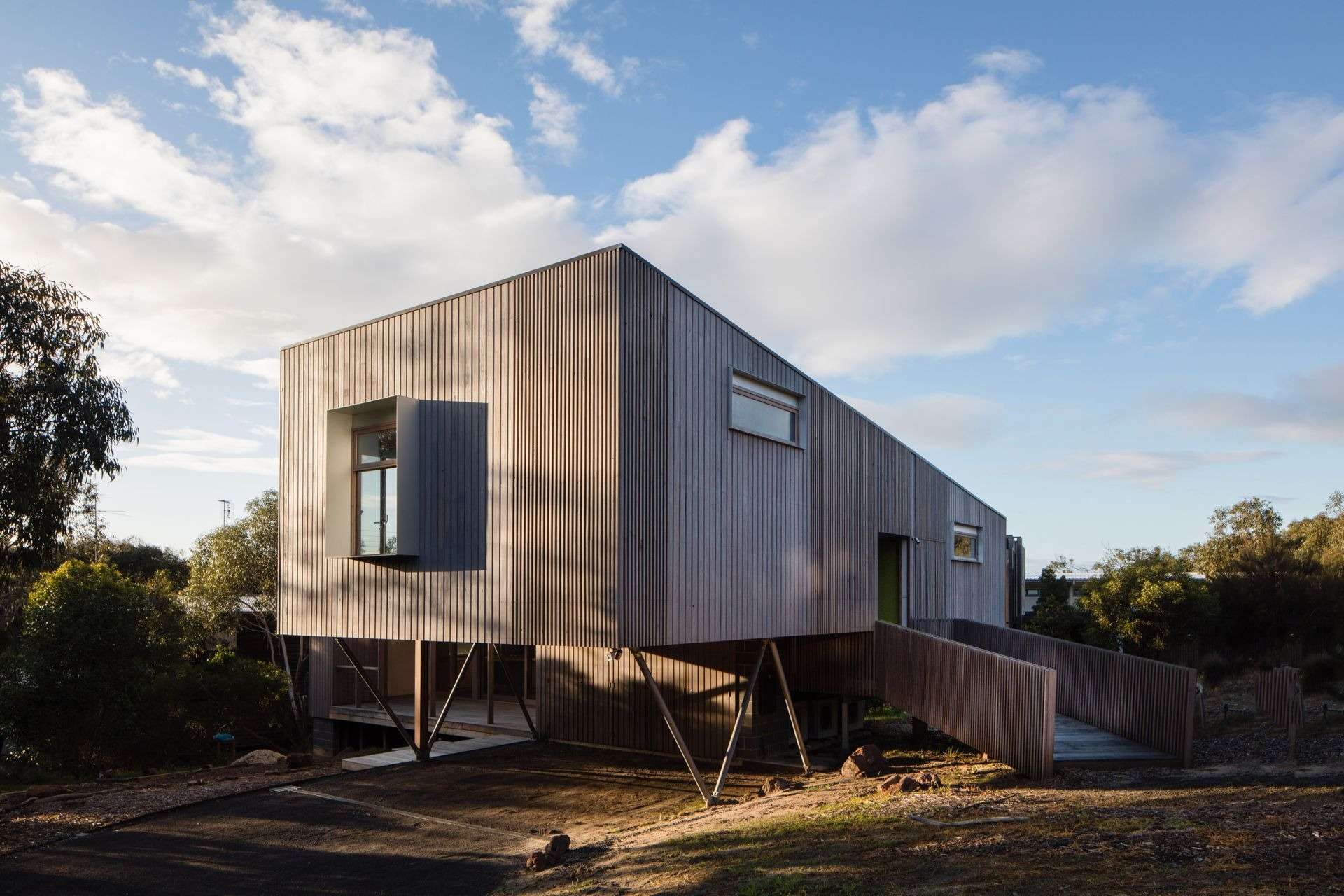 Elevated timber-clad home on slender angled supports, with a projecting window and sloping access ramp amid coastal vegetation.