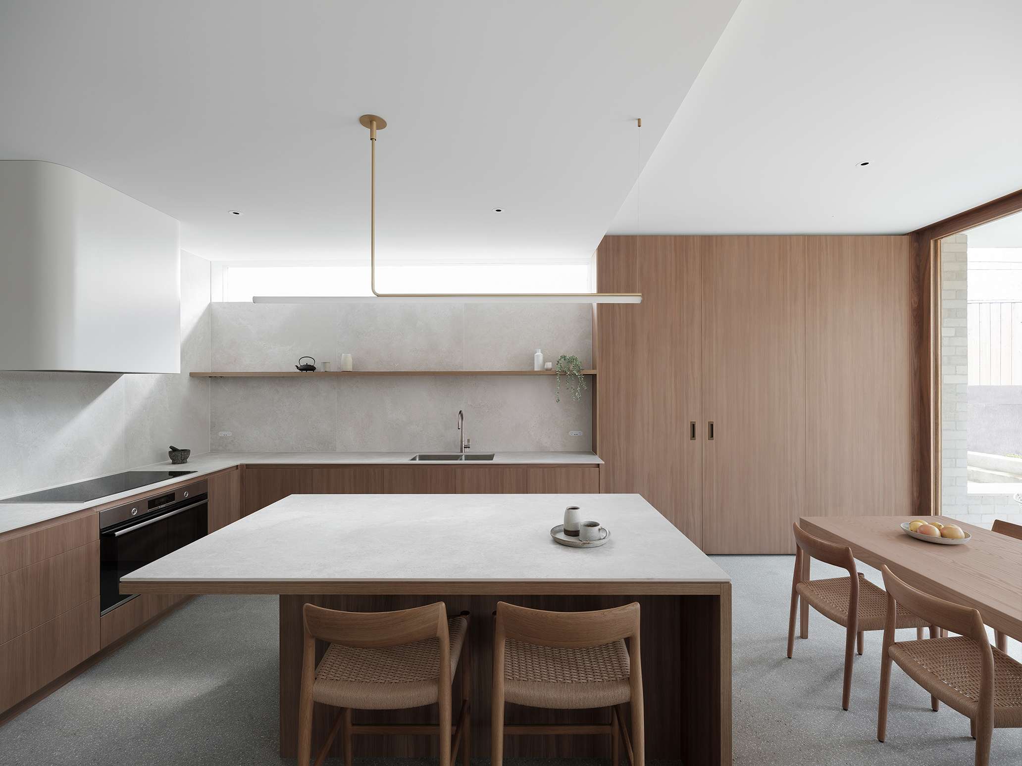 Bright kitchen with pale timber cabinetry, tall cupboards and a central island, with daylight entering through a high window and adjacent glazing.