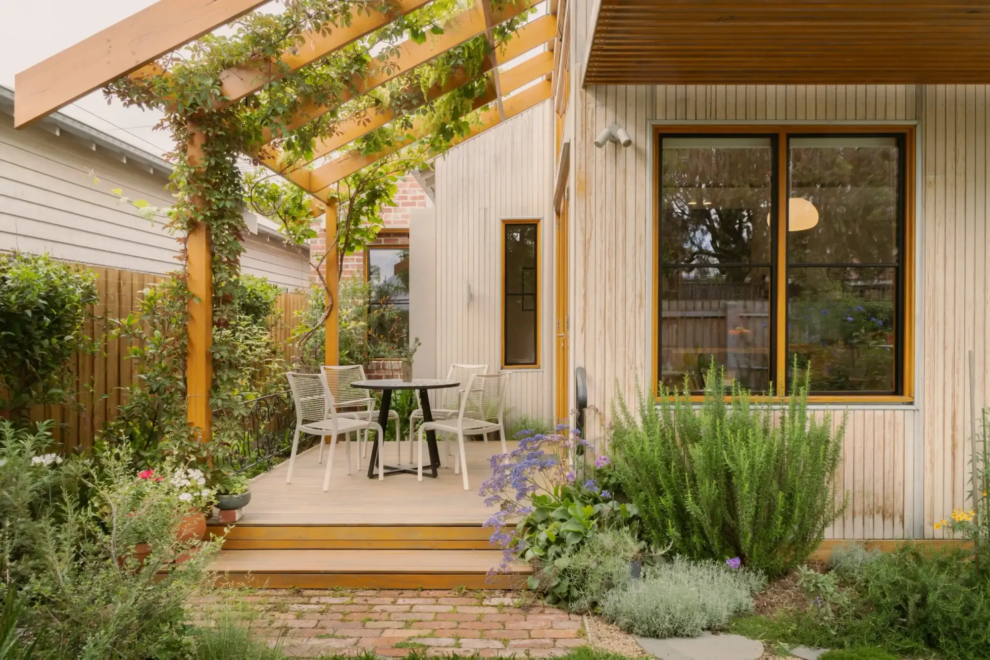 Timber deck with an outdoor dining setting beneath a vine-covered pergola, surrounded by garden planting beside a timber-clad home.