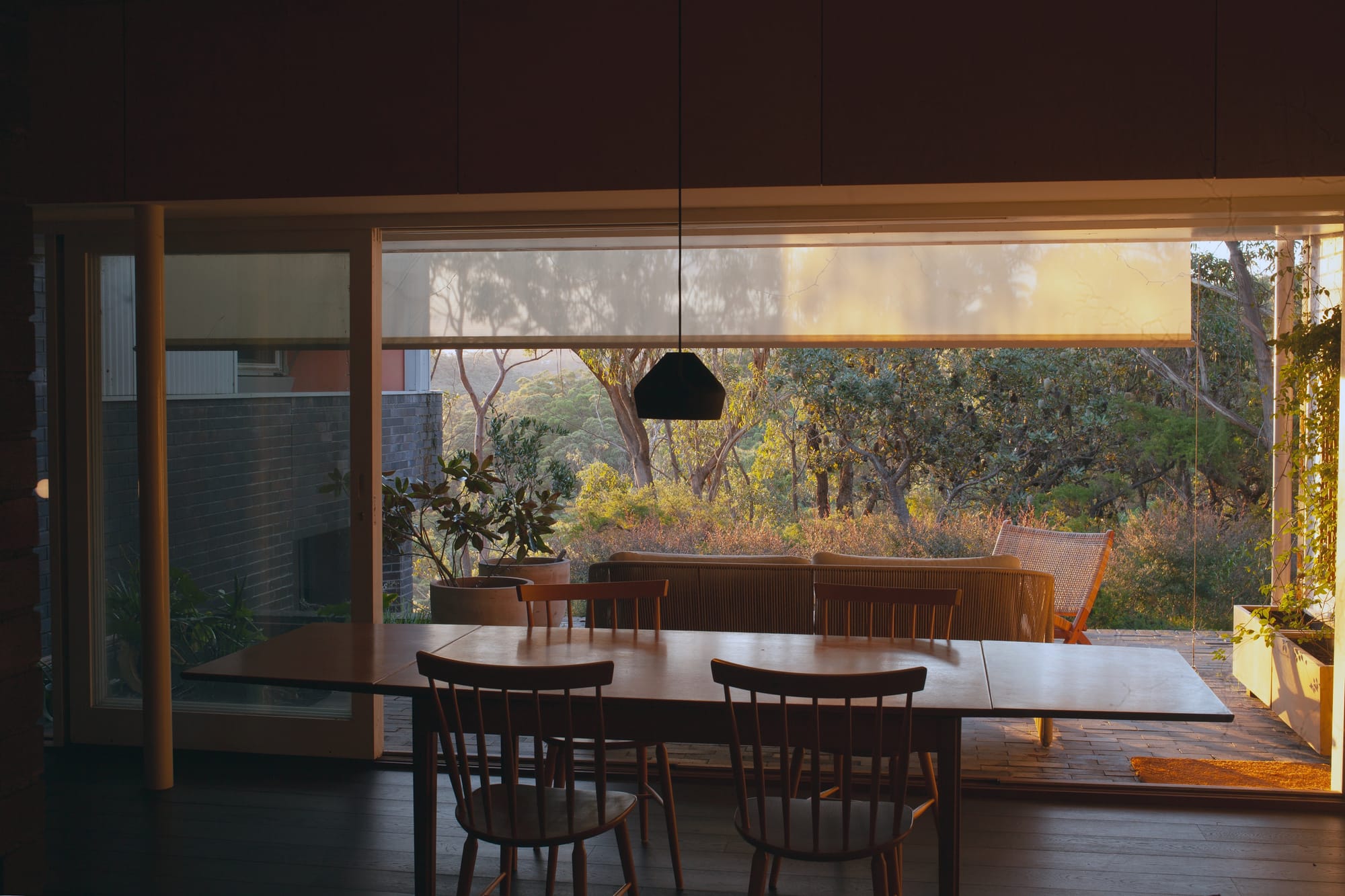 Warm sunlight falls across a terrace overlooking bushland, seen through broad glazing from a shaded dining room with a timber table, wooden chairs and a black pendant light.