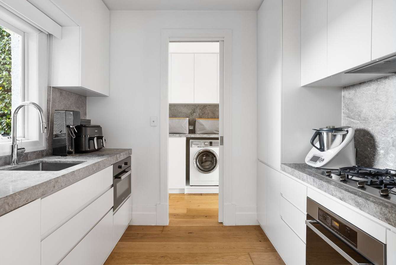 Galley kitchen with white cabinetry, grey benchtops, a sink beneath a window and timber flooring leading to an adjoining laundry.