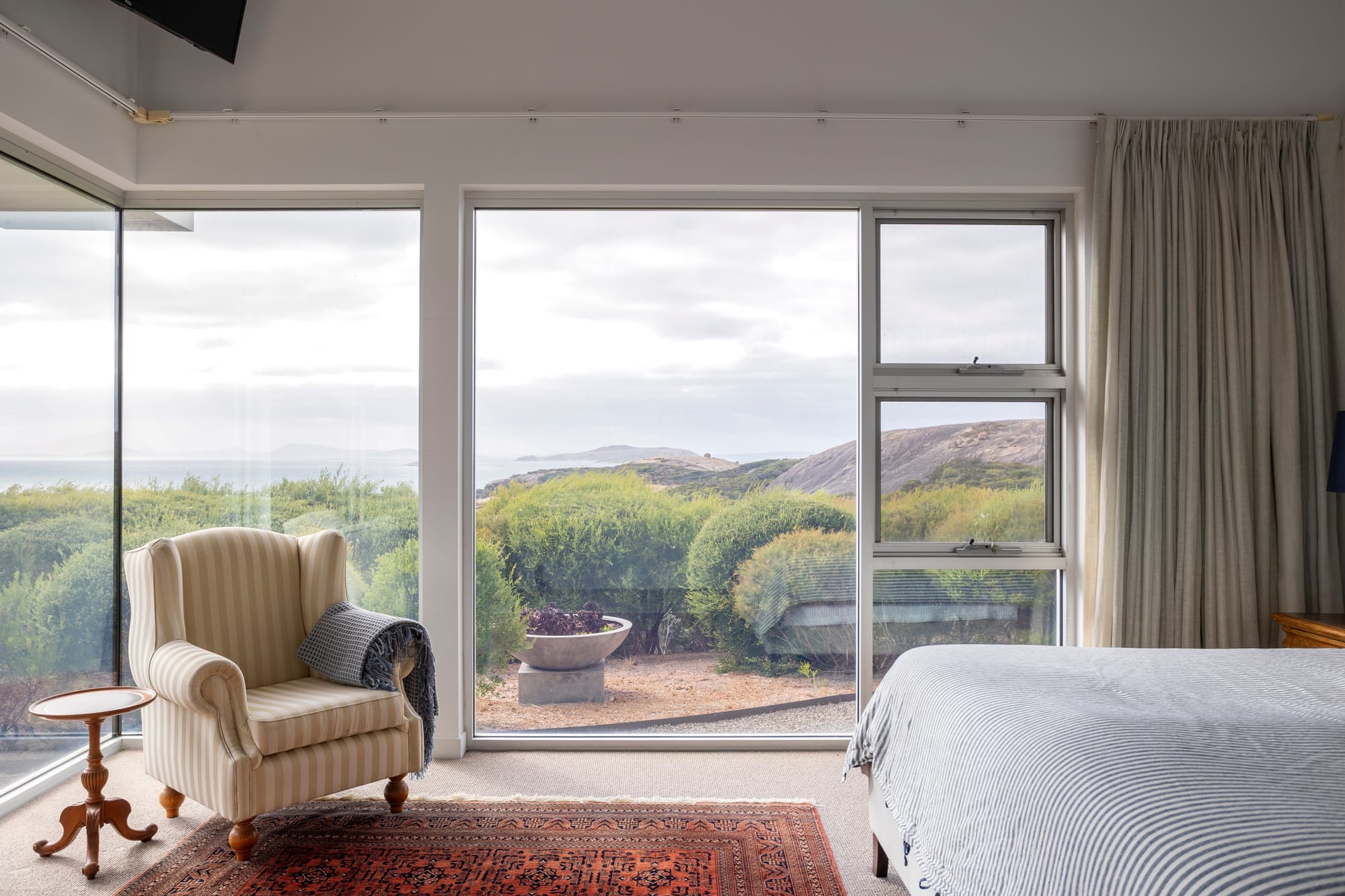 Large corner windows frame coastal vegetation, rocky outcrops and ocean views from a bedroom with a striped armchair, patterned rug and pale curtains.