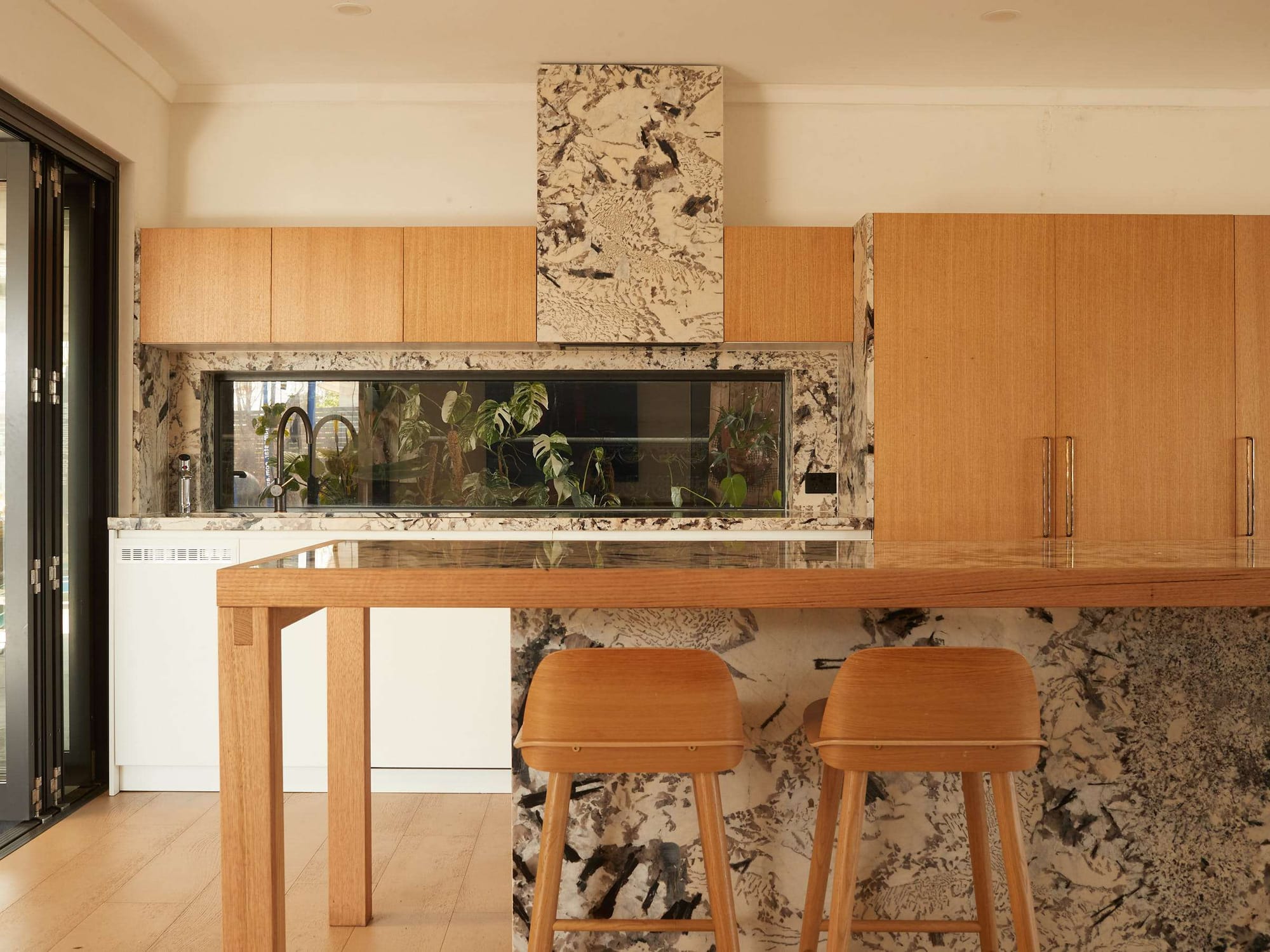 Kitchen with warm timber cabinetry, boldly patterned stone surfaces and a timber breakfast bar with two tan stools.