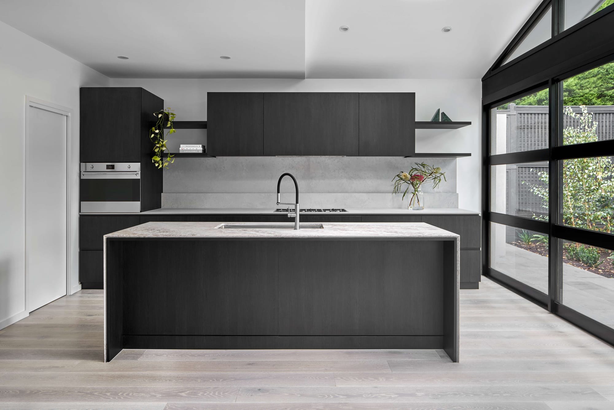 Kitchen with dark timber cabinetry, a stone island with an inset sink, pale timber flooring and tall windows overlooking the garden.