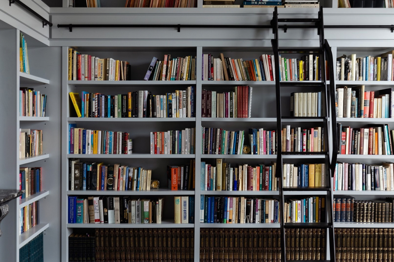 Floor-to-ceiling grey bookshelves filled with books, with a black library ladder mounted on a horizontal rail.