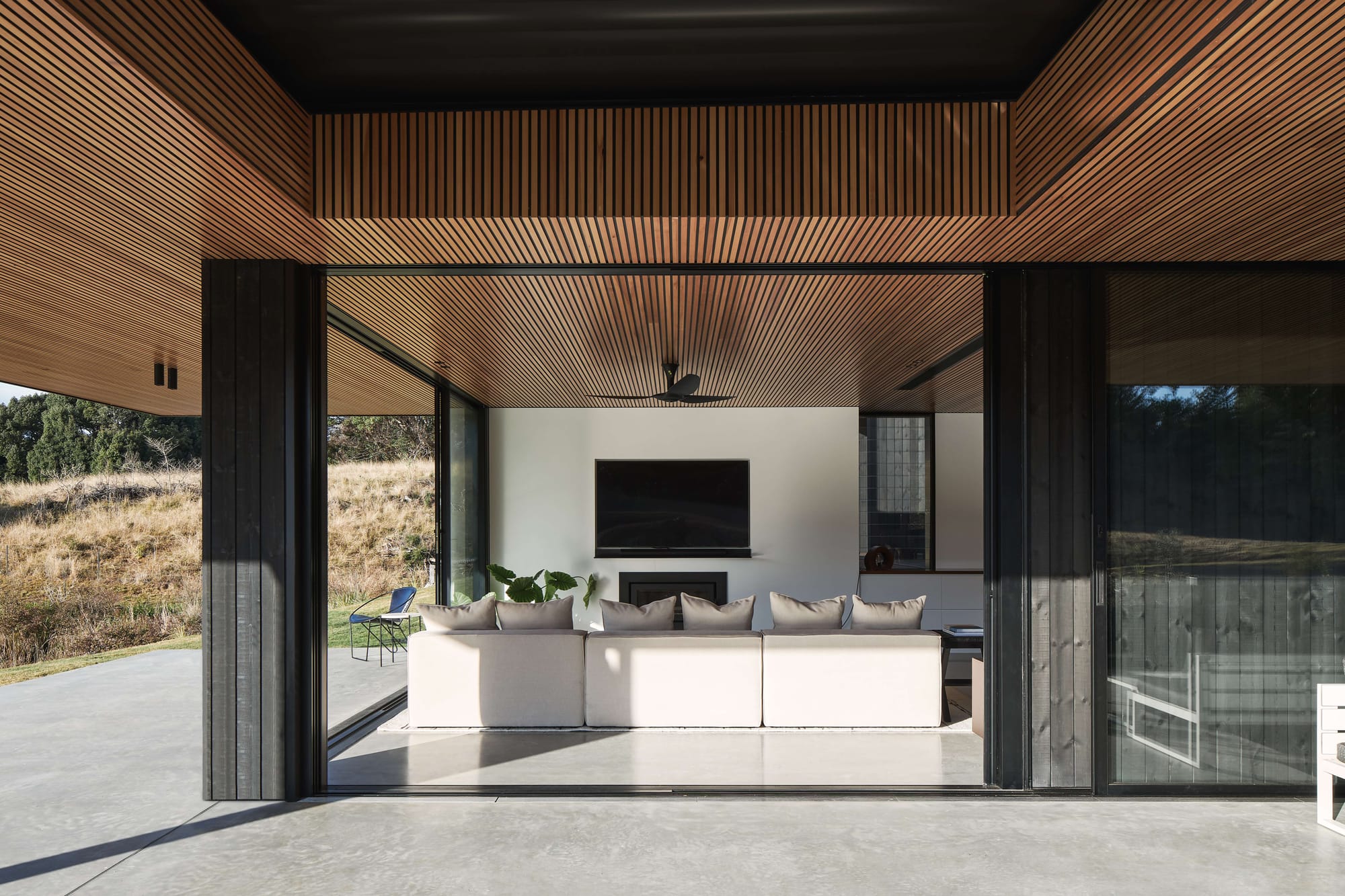 Sheltered terrace with timber batten ceilings and dark framed sliding glass doors opening to a living room overlooking the surrounding landscape.