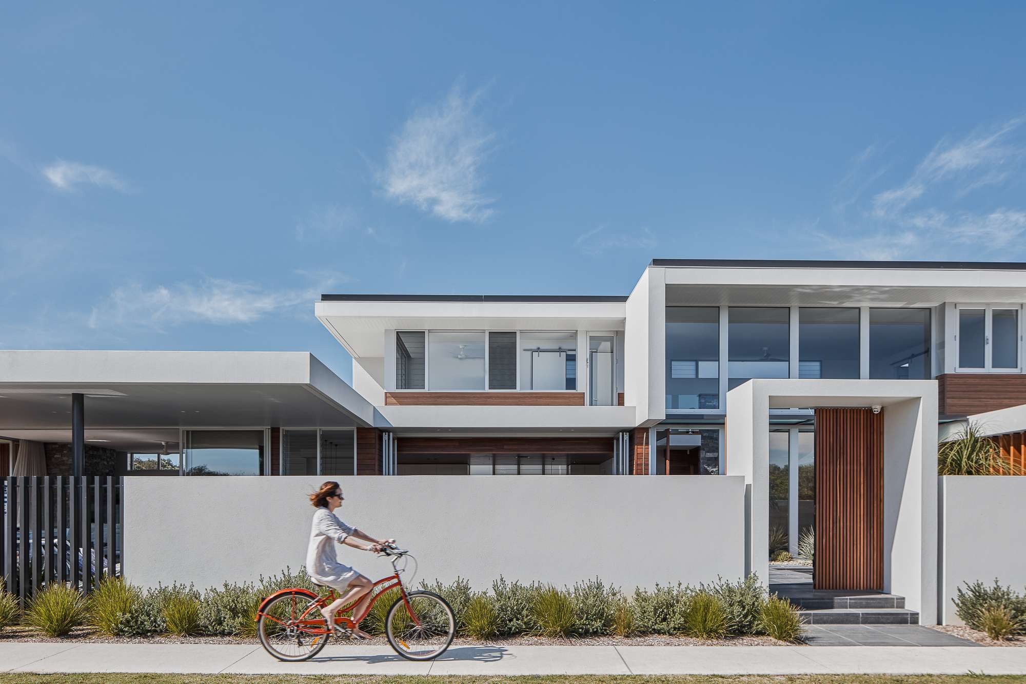 A cyclist passes a two storey coastal home with white walls, timber cladding, broad windows and projecting roof edges. A tall timber gate marks the entrance.