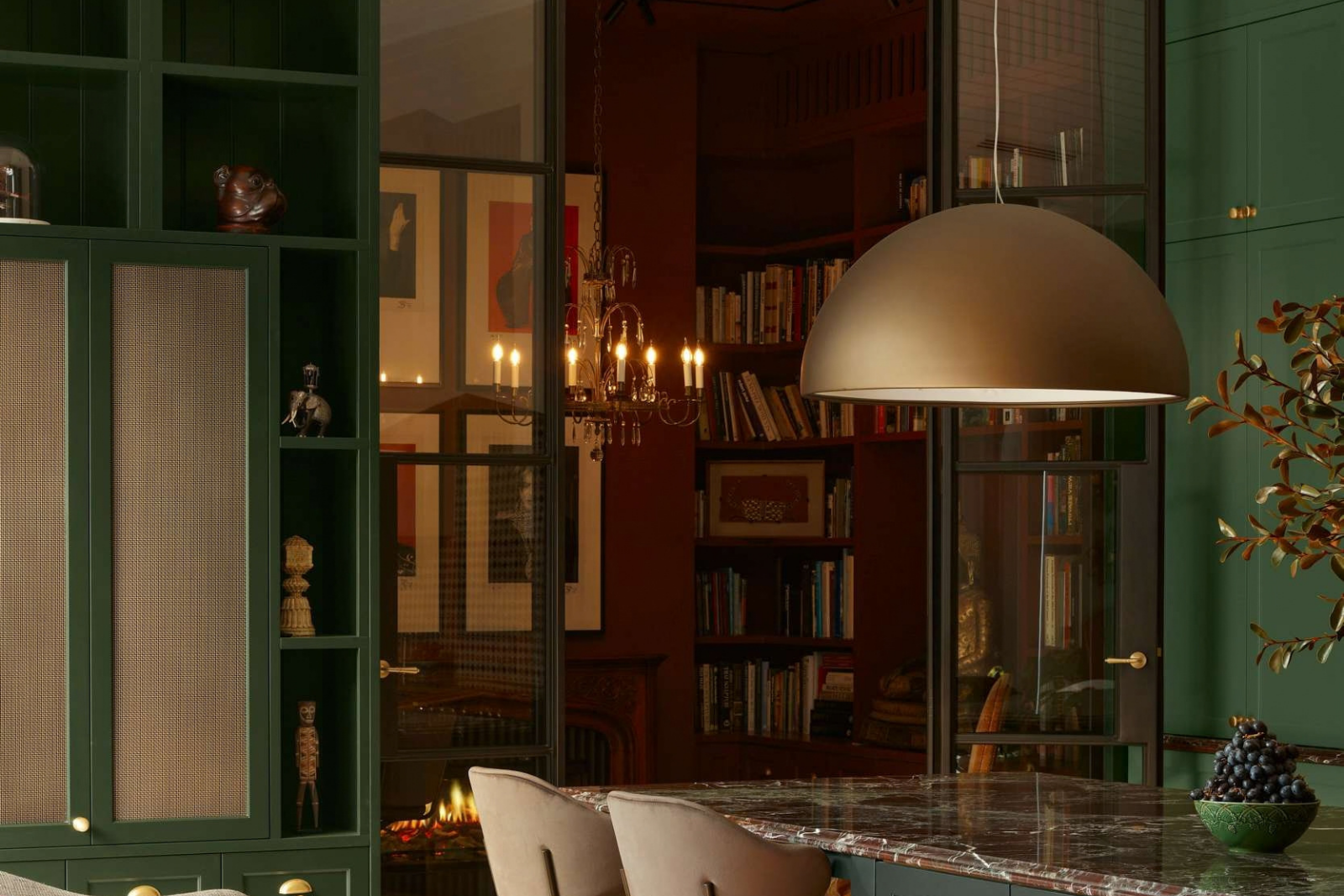 Deep green kitchen cabinetry and a stone island beneath a dome pendant, with glazed doors opening to a library with bookshelves, a chandelier and fireplace.