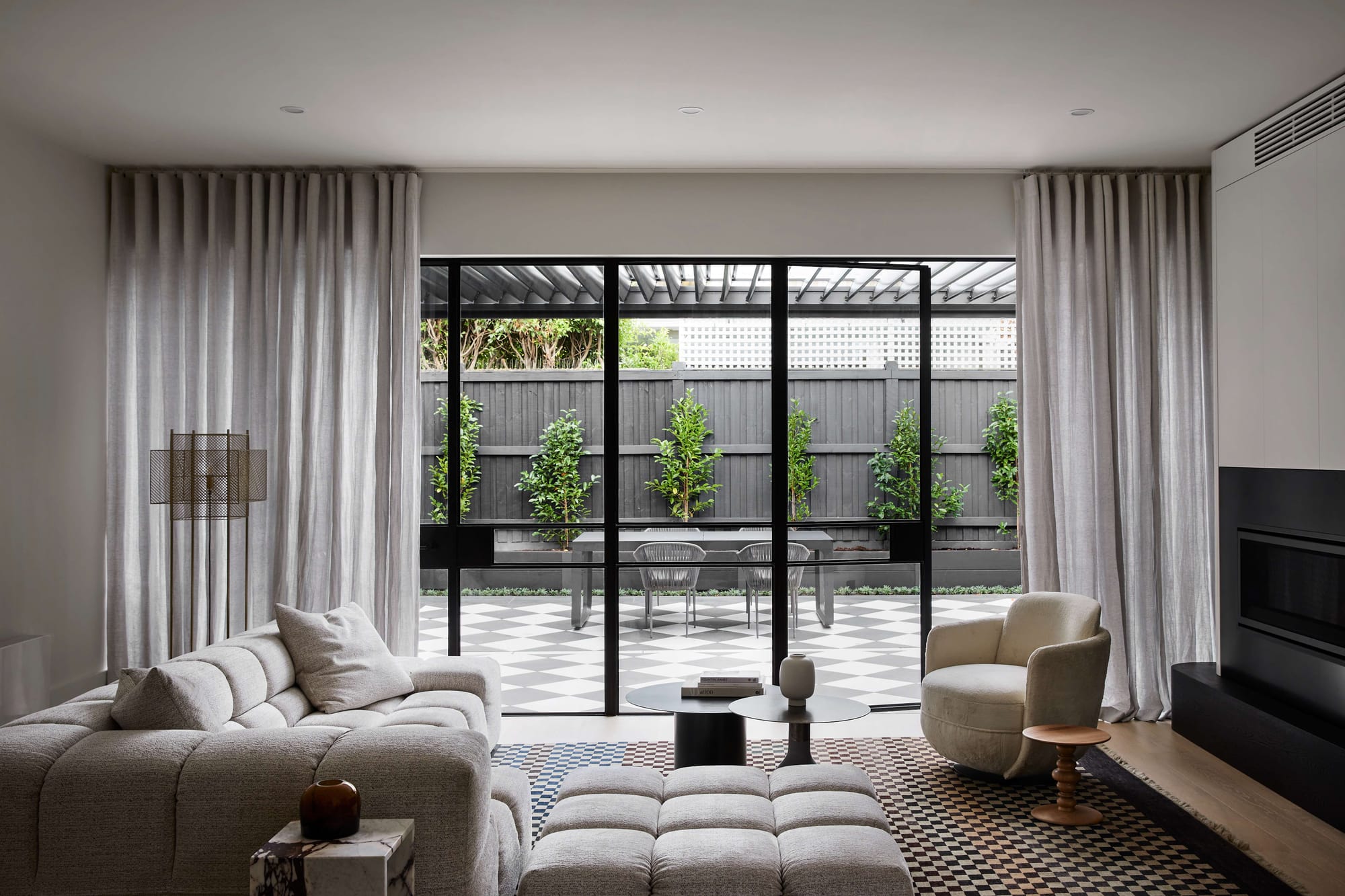 Living room with a grey sofa, upholstered ottoman and cream armchair, opening through black-framed glass doors to a checkerboard-paved courtyard.