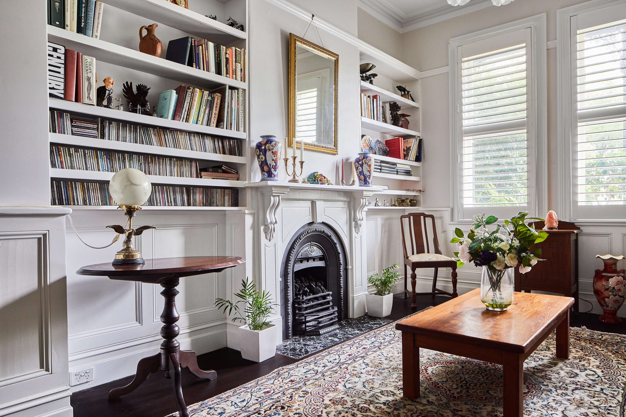 Traditional sitting room with white built-in bookshelves surrounding an ornate fireplace, plantation shutters, timber furniture and a patterned rug.