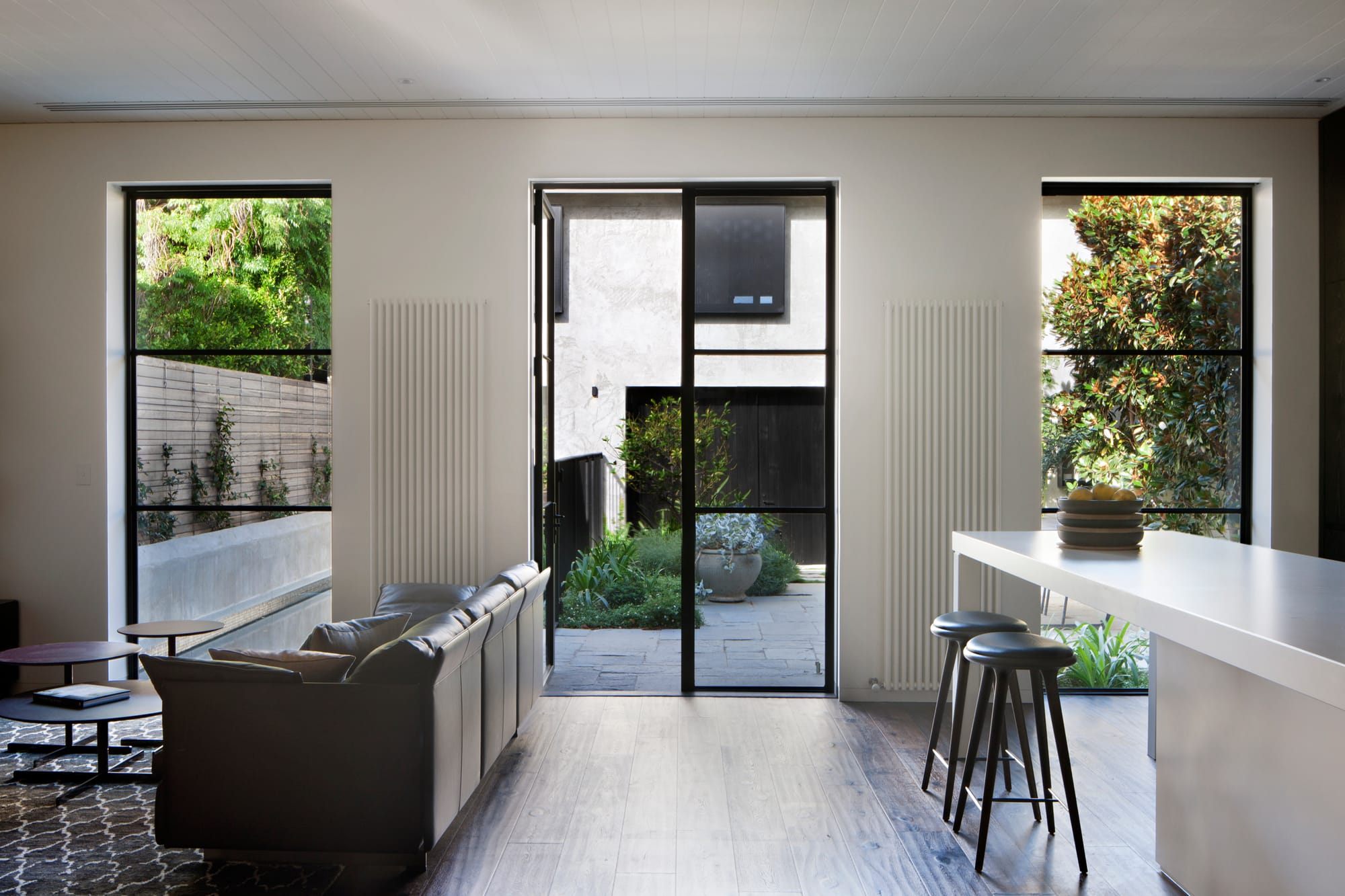 Open plan living and kitchen area with timber flooring, a grey sofa and white island, opening onto a planted courtyard through black steel framed glazing.