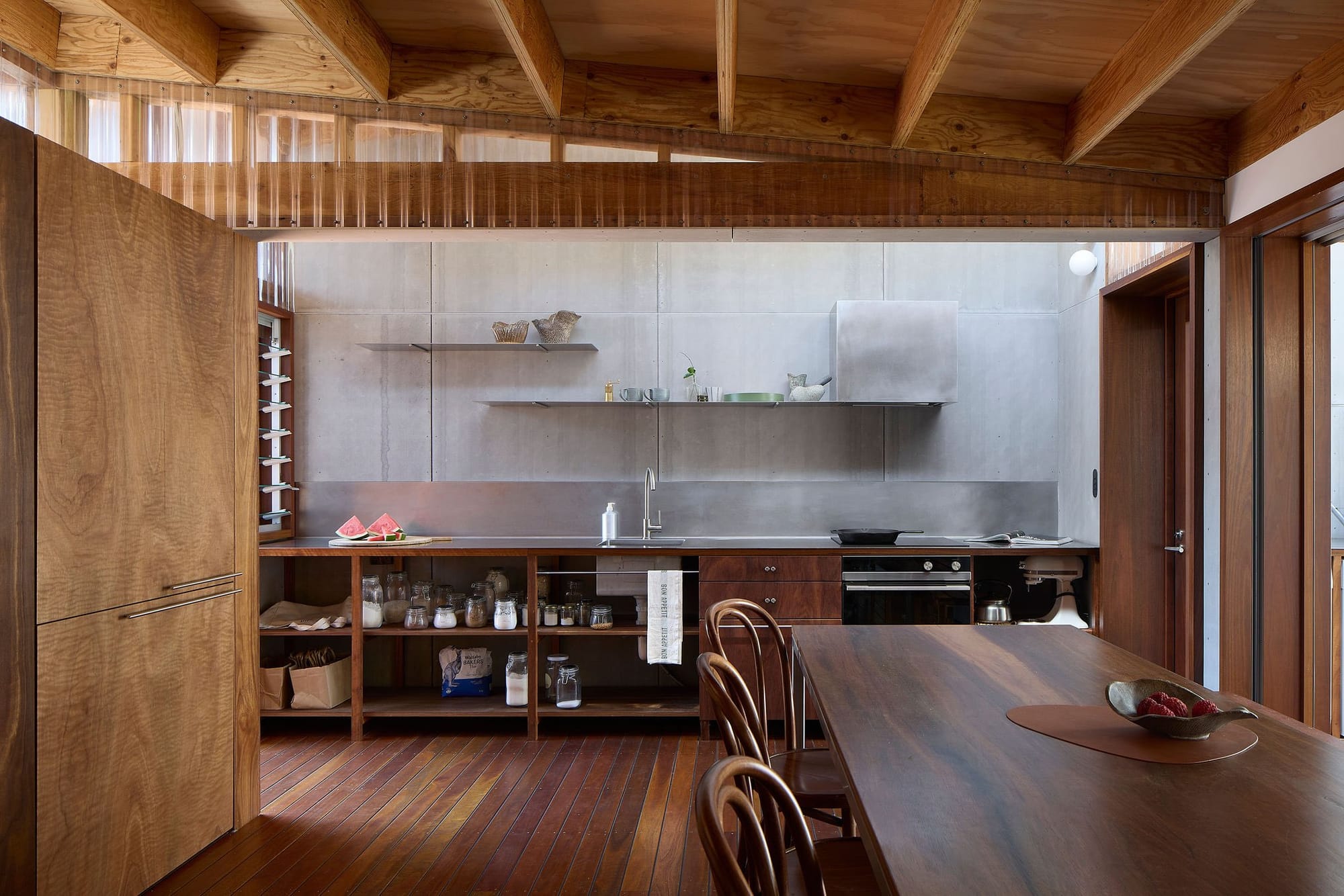 Timber kitchen and dining area at House that’s a Deck, with exposed ceiling beams, open shelving and a stainless steel benchtop illuminated by high windows.