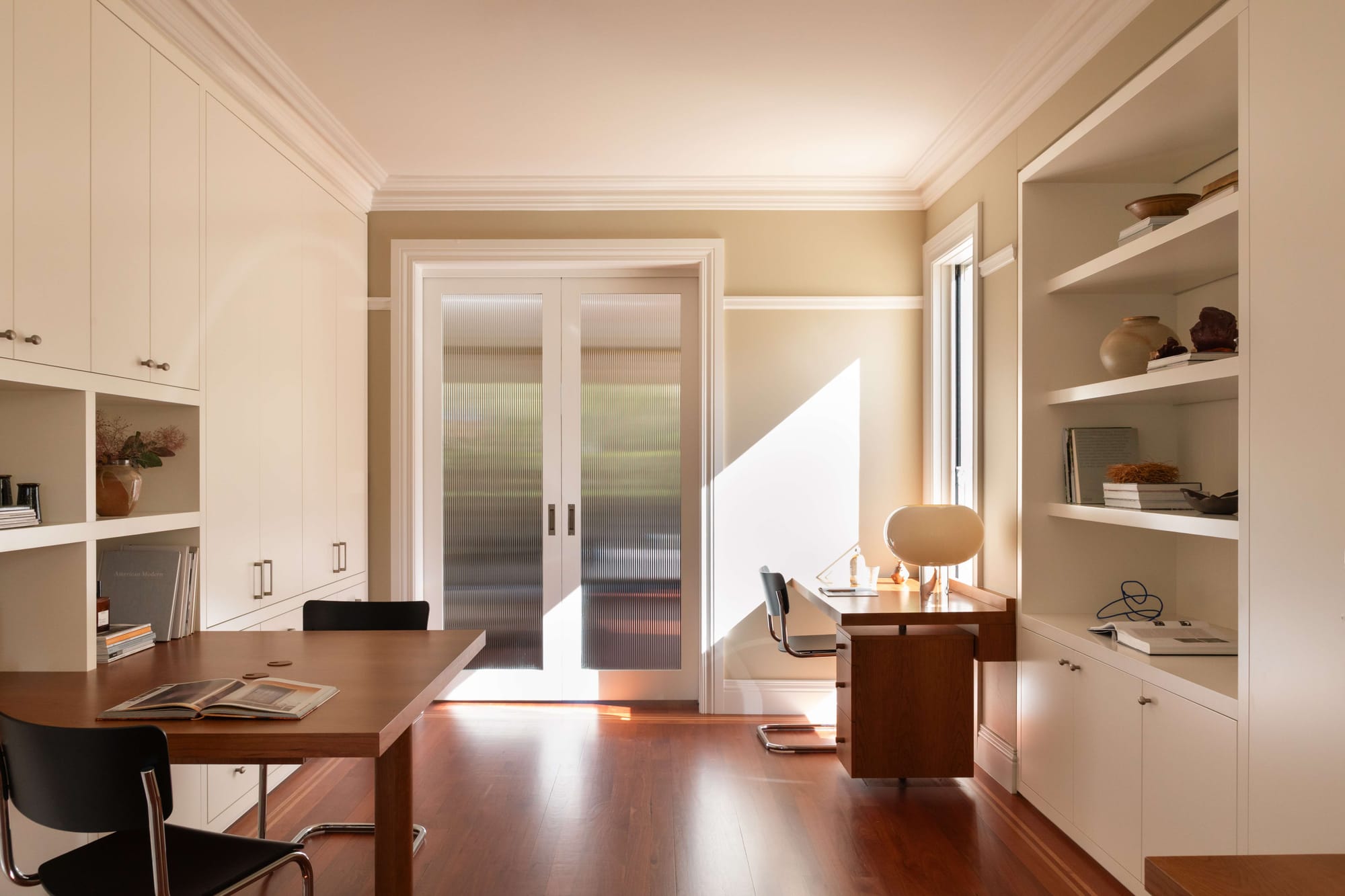 Sunlit study with cream built-in cupboards and shelves, a timber worktable, a separate desk beside the window and reeded glass double doors.